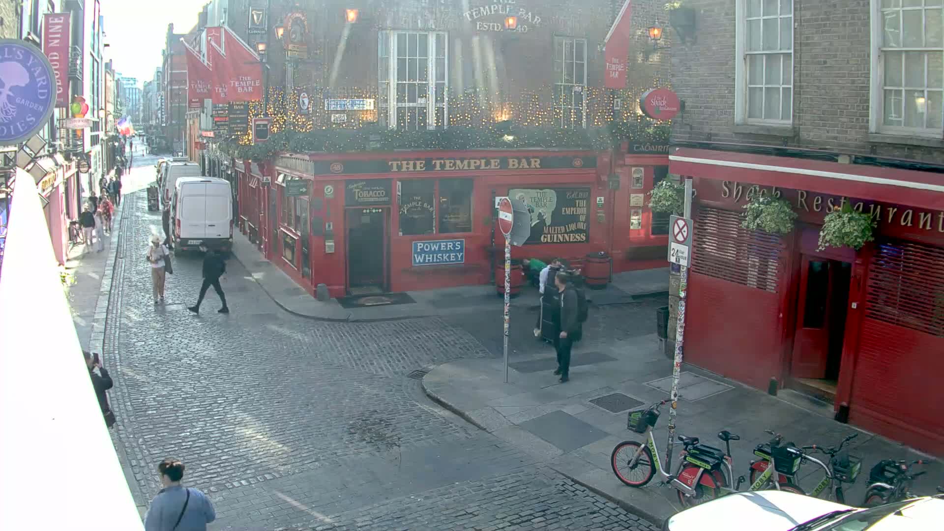 A cobblestone street scene on a sunny day shows pedestrians walking past red brick buildings, including a pub and restaurant, with several bicycles parked nearby.
