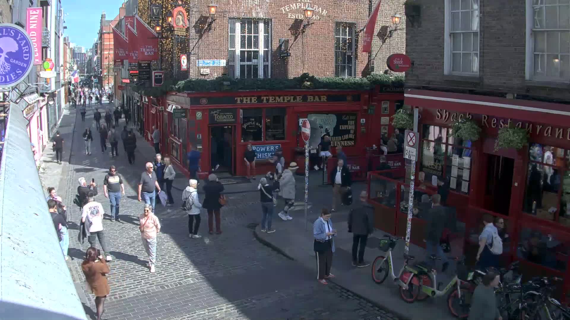 A sunny day on a cobblestone street in a city, with many people walking past red brick buildings and pubs.