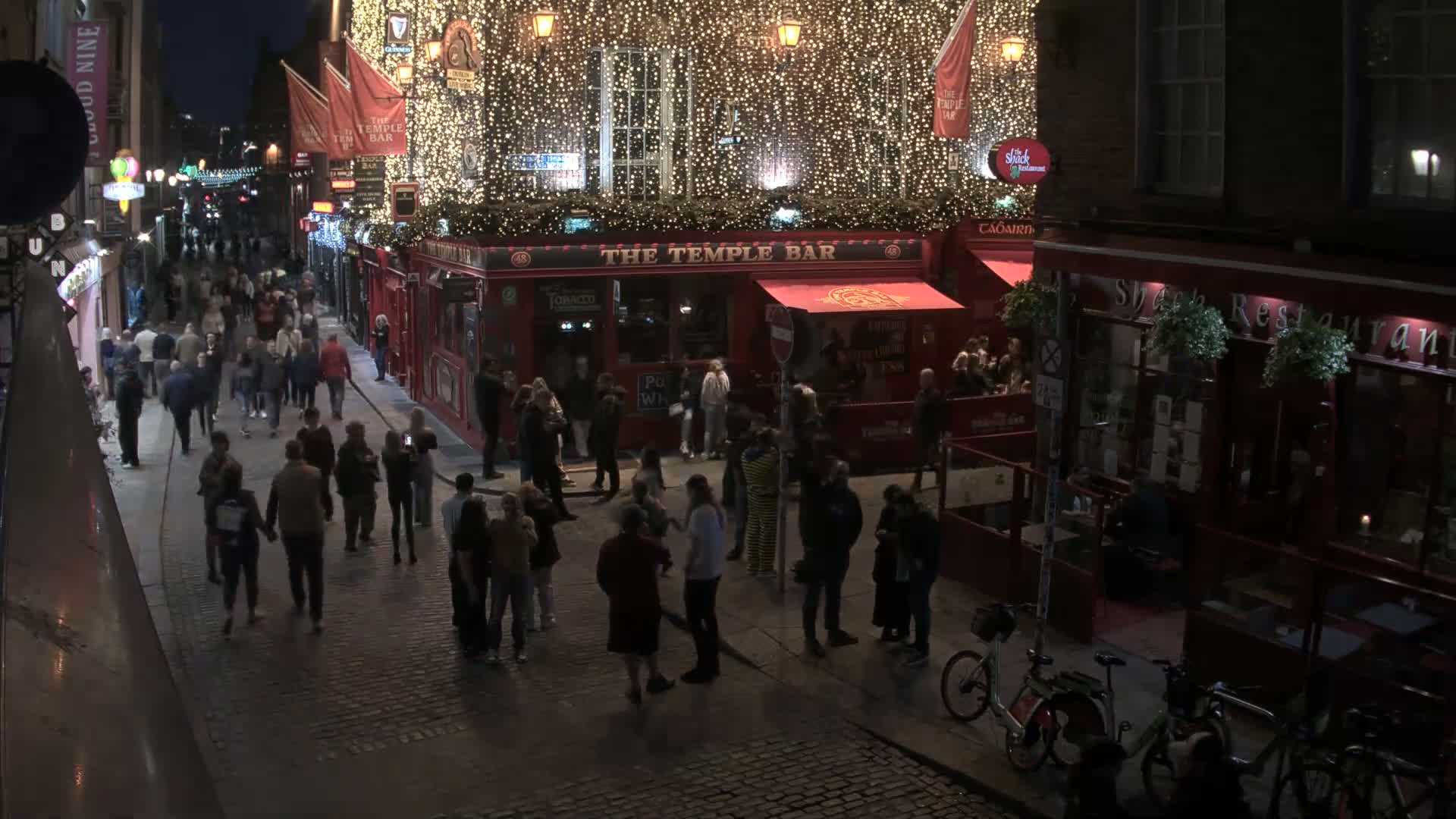 A nighttime street scene shows a cobblestone street crowded with people walking past illuminated pubs and restaurants decorated with string lights.