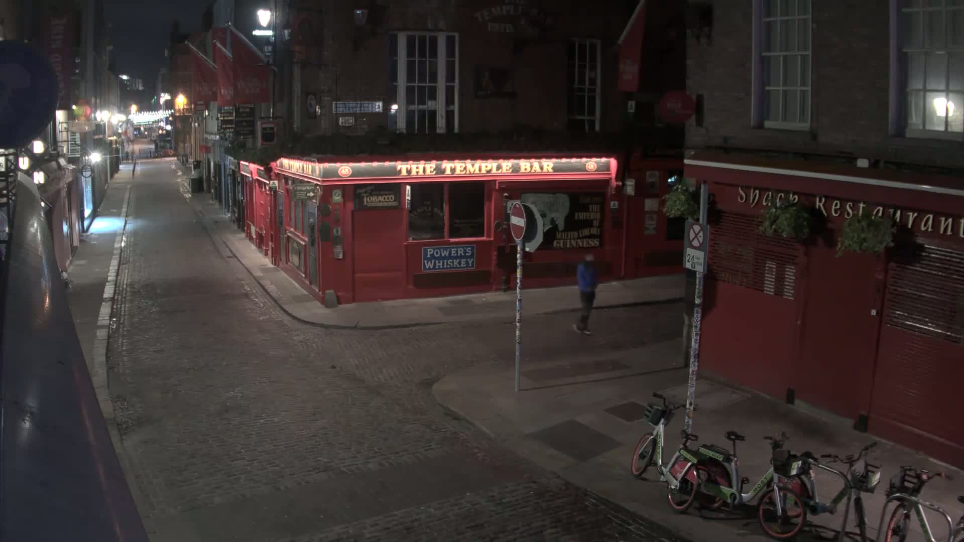 A nighttime, cobblestone street scene shows a red pub called "The Temple Bar" and several parked bicycles, with a single pedestrian walking past.