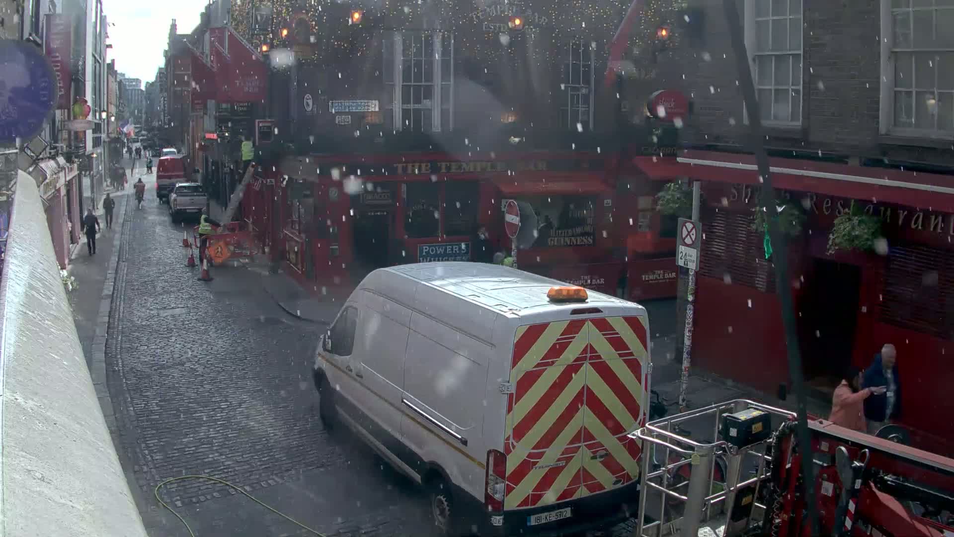 A narrow cobblestone street in the snow, lined with red brick buildings, with a white van and some pedestrians visible.