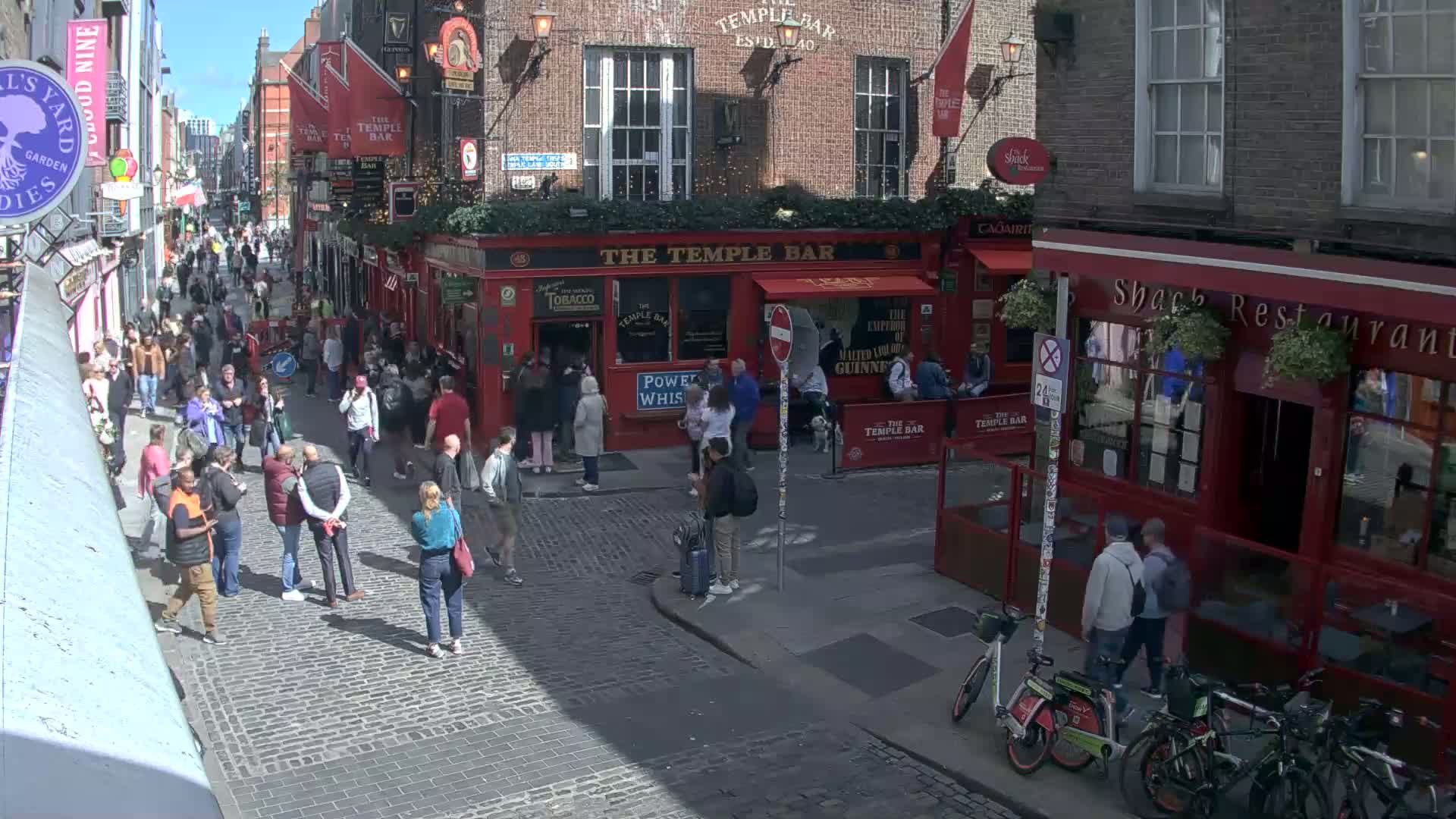 A sunny day on a cobblestone street with many people walking past red brick buildings and pubs.
