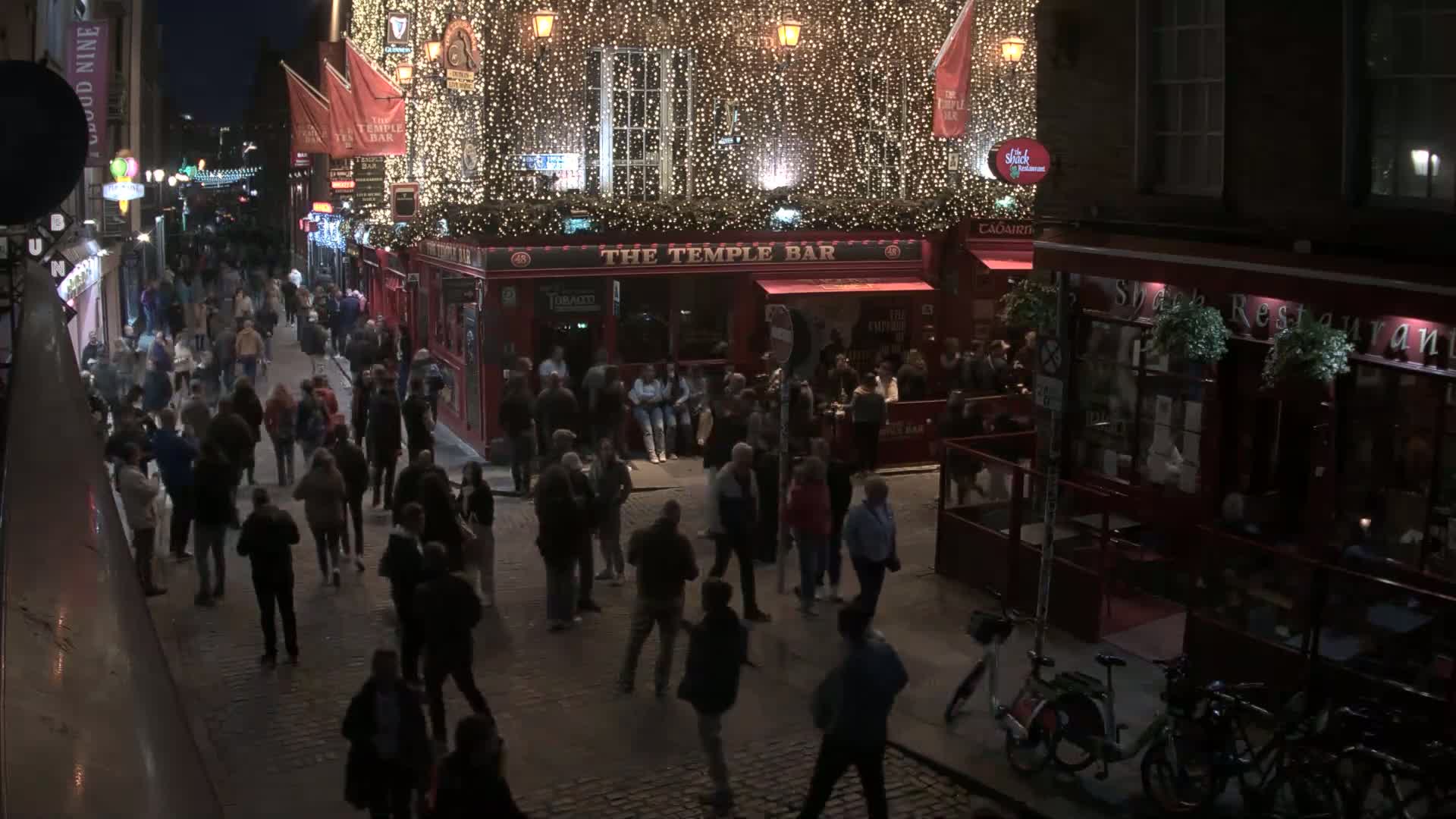 A nighttime, crowded cobblestone street scene features a brightly lit pub adorned with string lights, surrounded by pedestrians and a few bicycles parked nearby.
