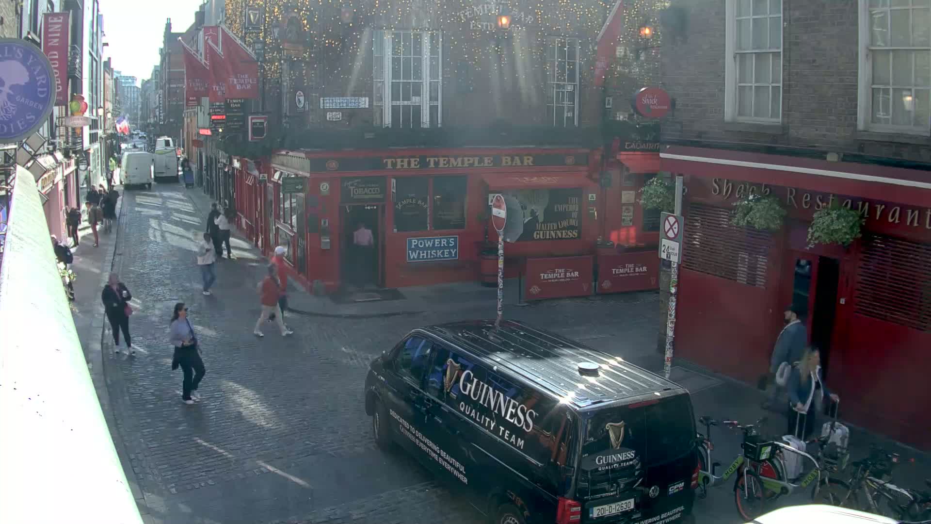 A sunny day on a cobblestone street in a city, with pedestrians walking past red brick buildings and a black Guinness van parked nearby.