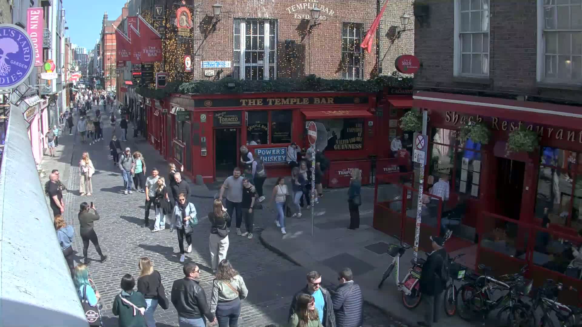 A sunny day on a cobblestone street, crowded with pedestrians walking past red brick buildings with pubs and restaurants.