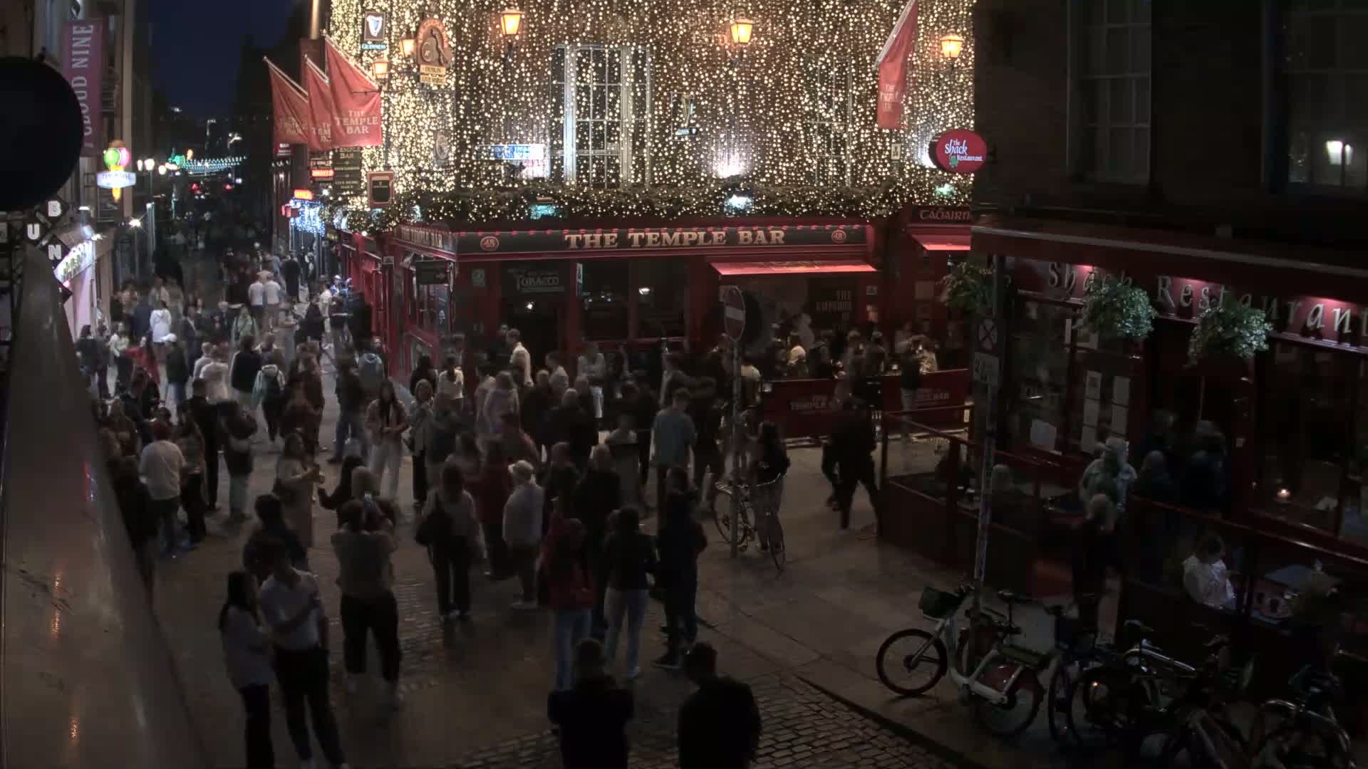 A nighttime, crowded cobblestone street scene shows many people walking around numerous pubs and restaurants decorated with string lights.