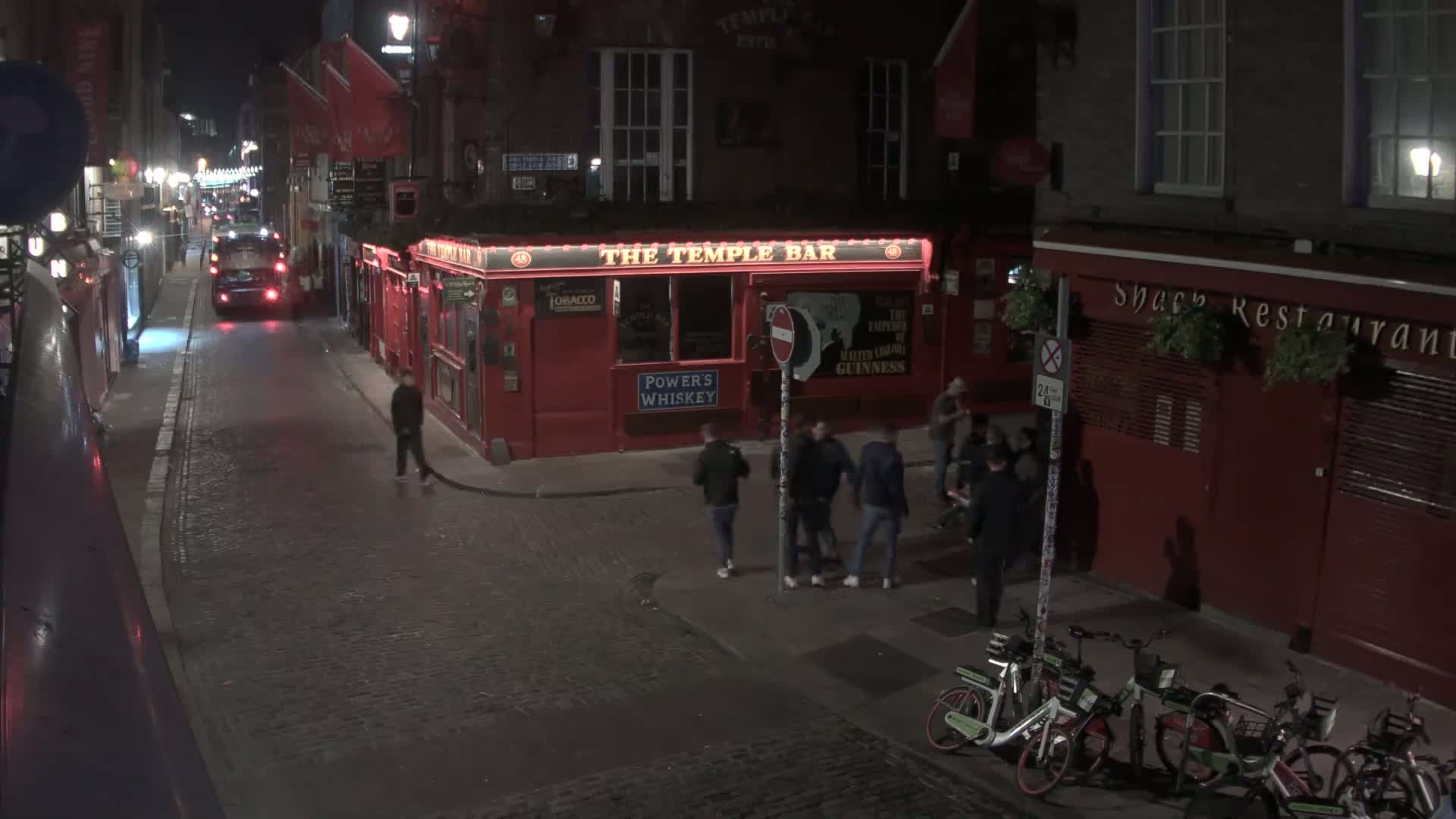 A nighttime street scene shows a cobblestone road with several people walking near a red building housing a bar and restaurant, with bicycles parked nearby.