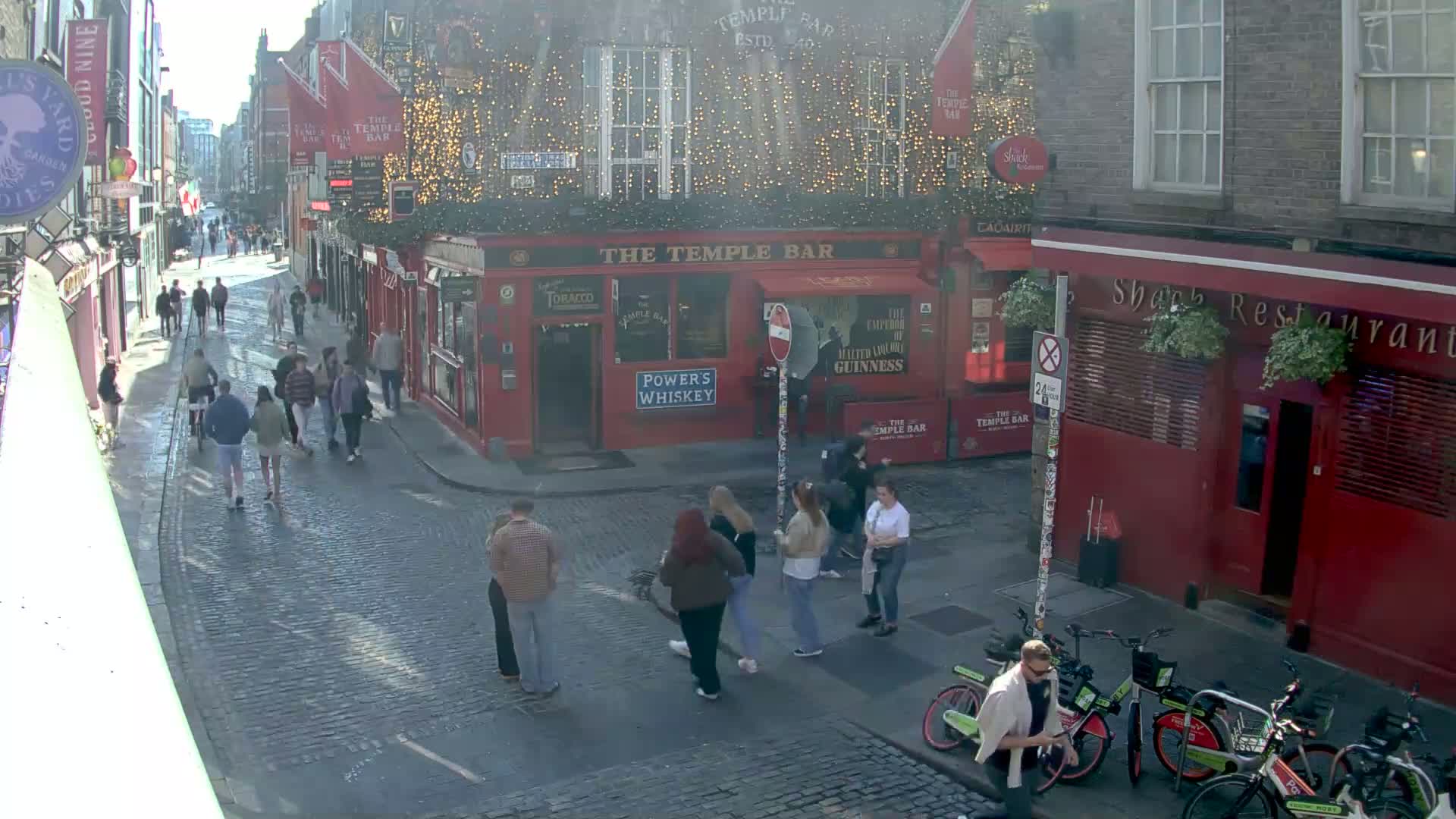A cobblestone street scene shows pedestrians walking past red brick buildings, including a pub and restaurant, on a sunny day.
