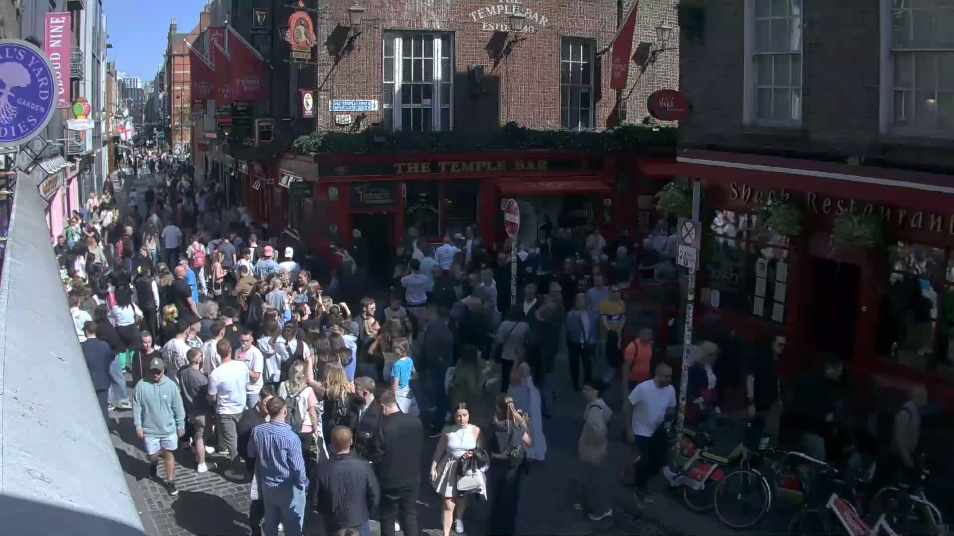 A large crowd of people is walking on a sunny day along a cobblestone street lined with brick buildings and pubs.