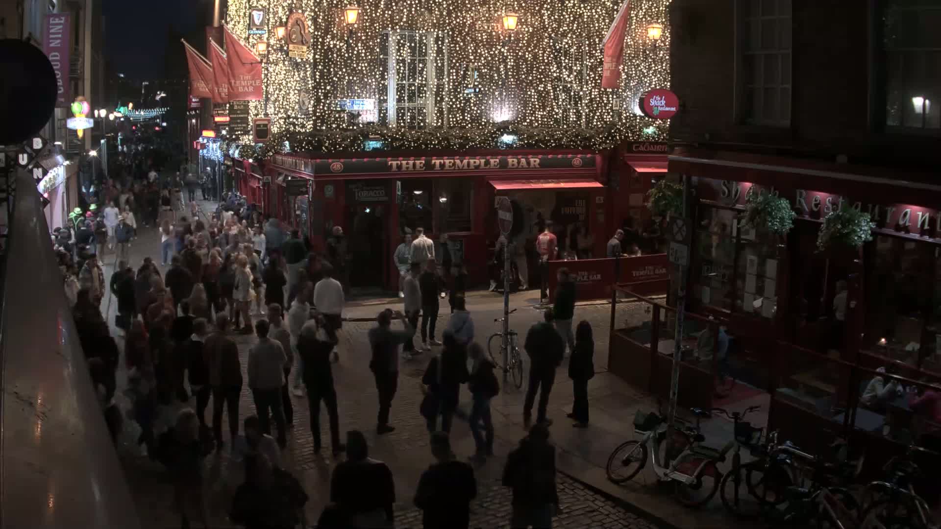 A nighttime street scene shows a large crowd of people gathered outside of a brightly lit pub decorated with string lights.