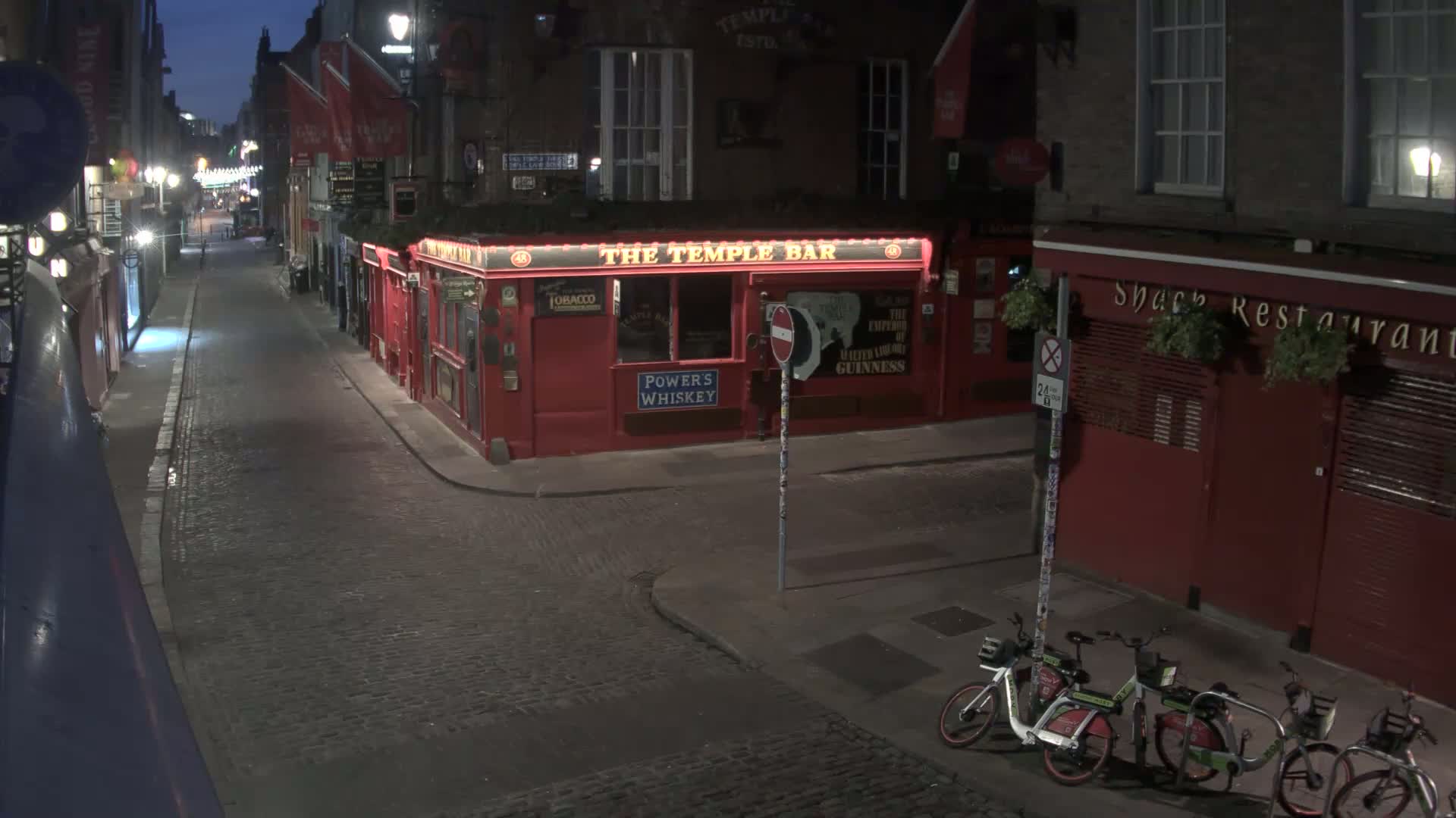 A dark, cobblestone street at night shows a red building with "The Temple Bar" signage, and two parked bicycles; other red buildings line the street.