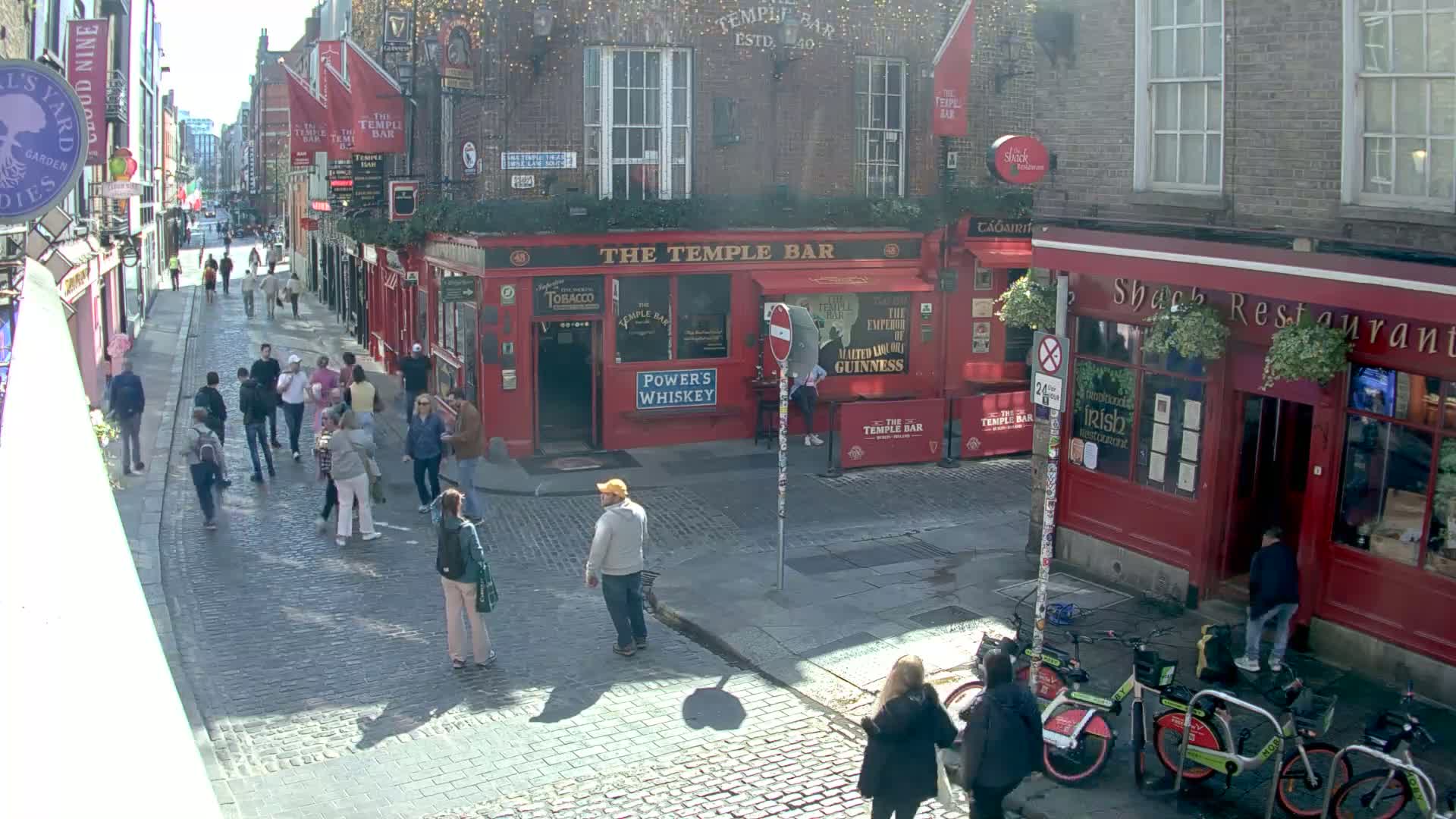 A sunny cobblestone street in a city, lined with red brick buildings, including a pub and a restaurant, with several pedestrians and bicycles.