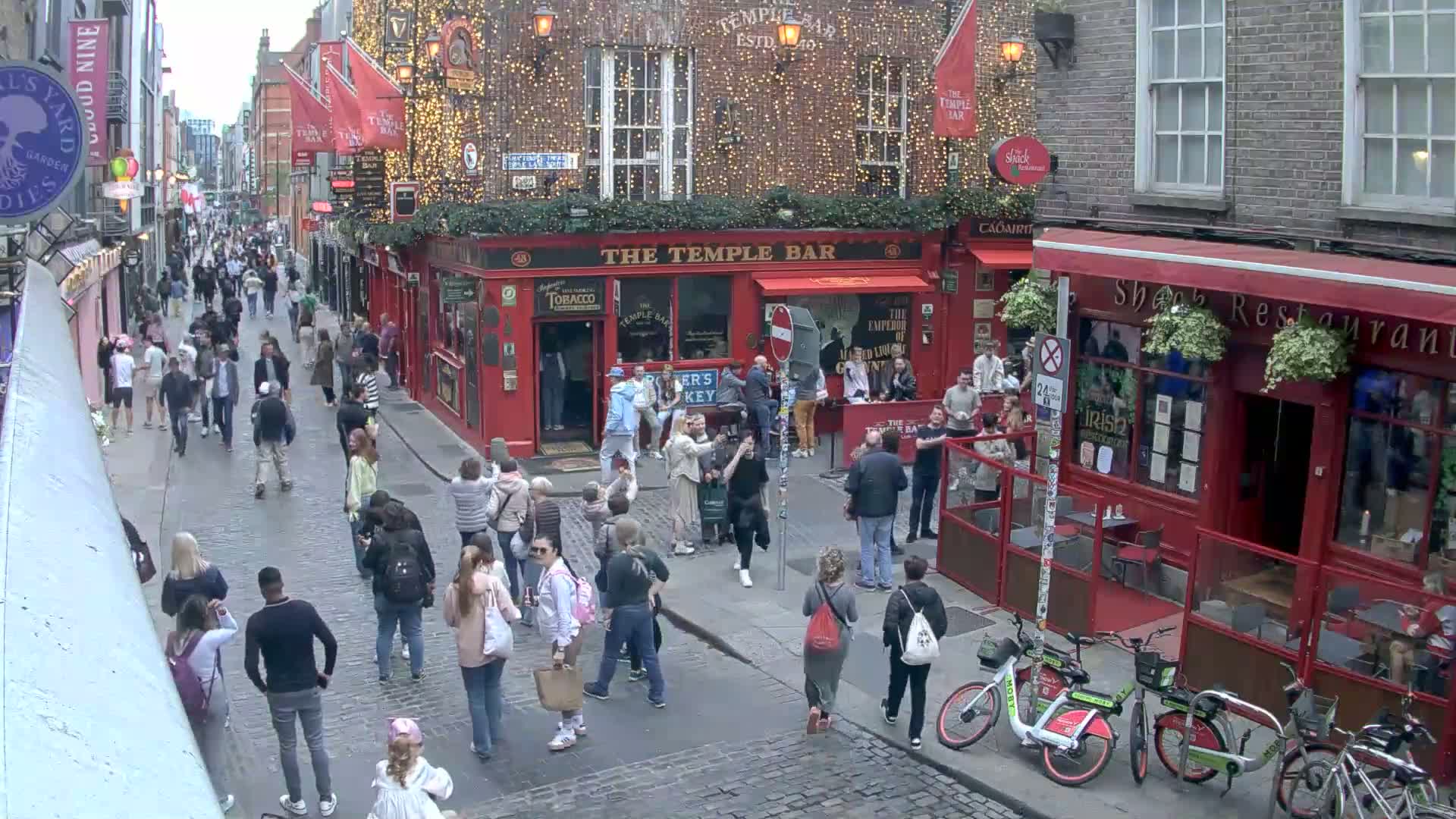 A bustling cobblestone street, under an overcast sky, is filled with pedestrians walking past red-brick buildings, including a pub with string lights.