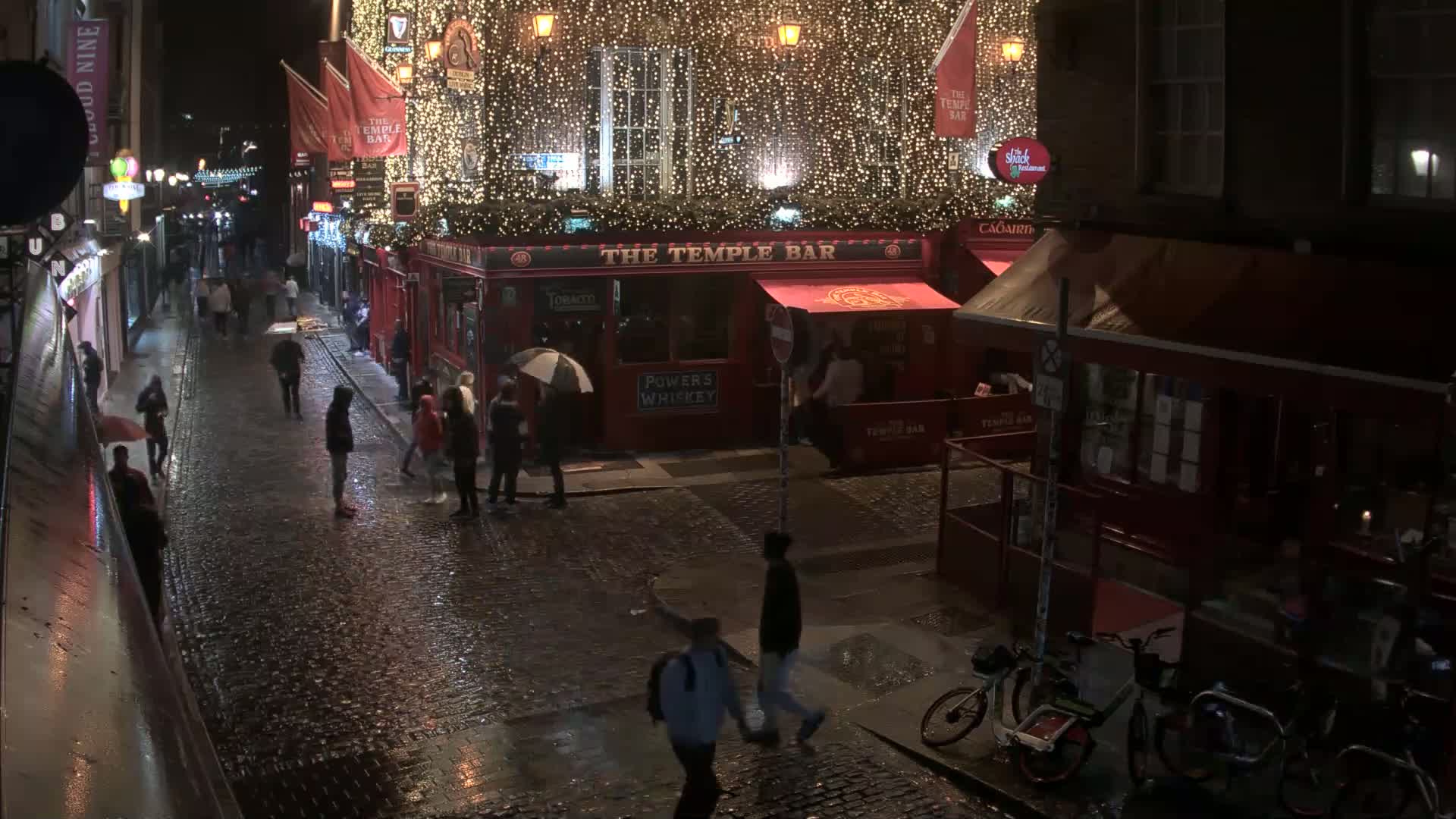 A rain-slicked cobblestone street at night, populated by pedestrians and a few bicycles, features a brightly lit pub adorned with string lights.