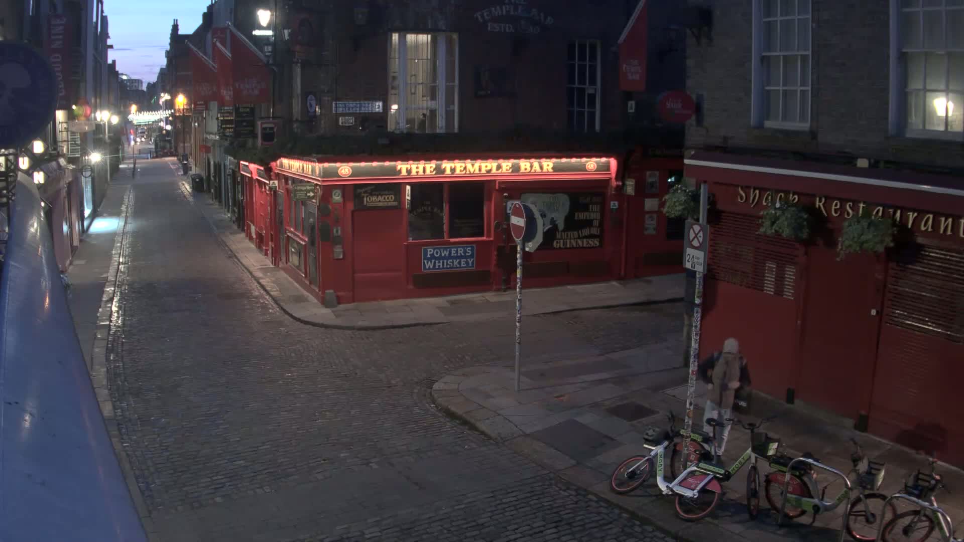 A cobblestone street at night, dimly lit, shows a red pub and restaurant with bicycles parked nearby under a twilight sky.