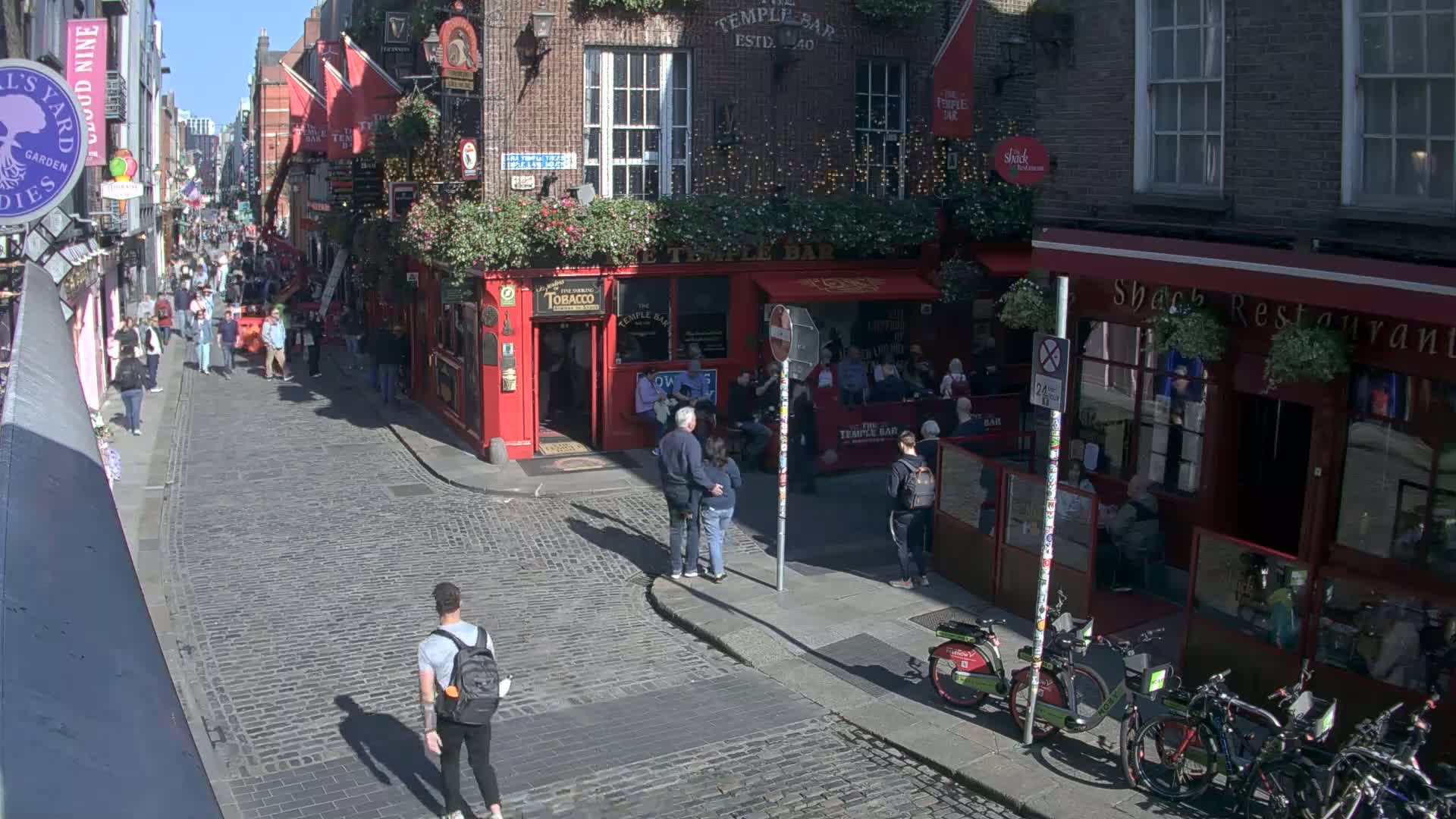 A sunny cobblestone street is filled with pedestrians and several red-brick buildings with outdoor seating areas.