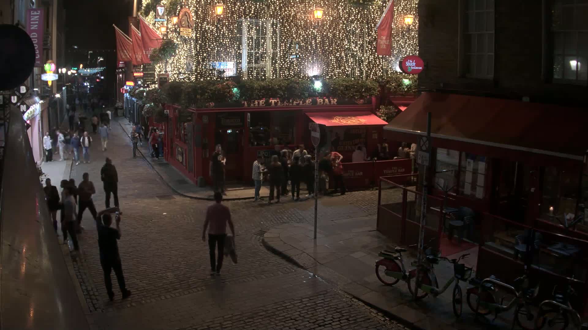 A nighttime street scene shows a cobblestone street with people walking past a brightly lit bar decorated with string lights, and some bicycles parked nearby.