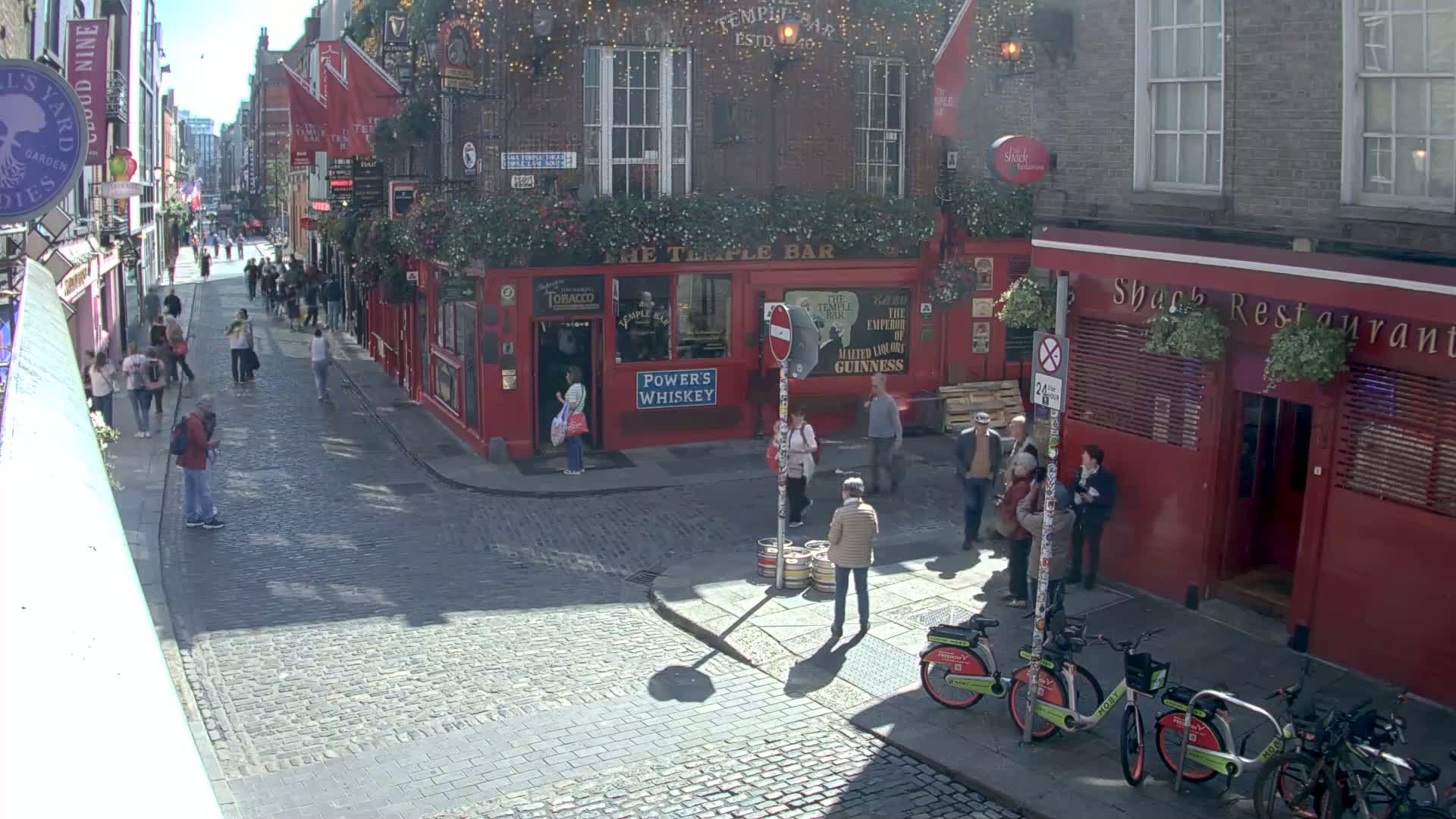 A sunny cobblestone street scene shows pedestrians walking past red brick buildings, one of which is a pub, with several bicycles parked nearby.