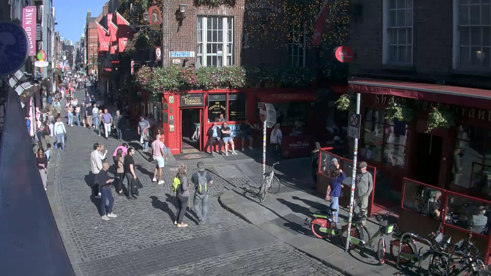 A cobblestone street is crowded with pedestrians on a sunny day, lined with buildings, including pubs and restaurants, and a few bicycles are parked along the side.