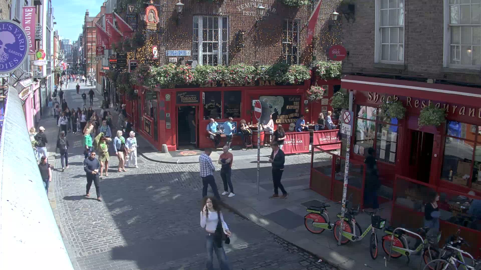 A cobblestone street in bright sunlight, bustling with pedestrians, features a red-brick pub with outdoor seating and a restaurant.