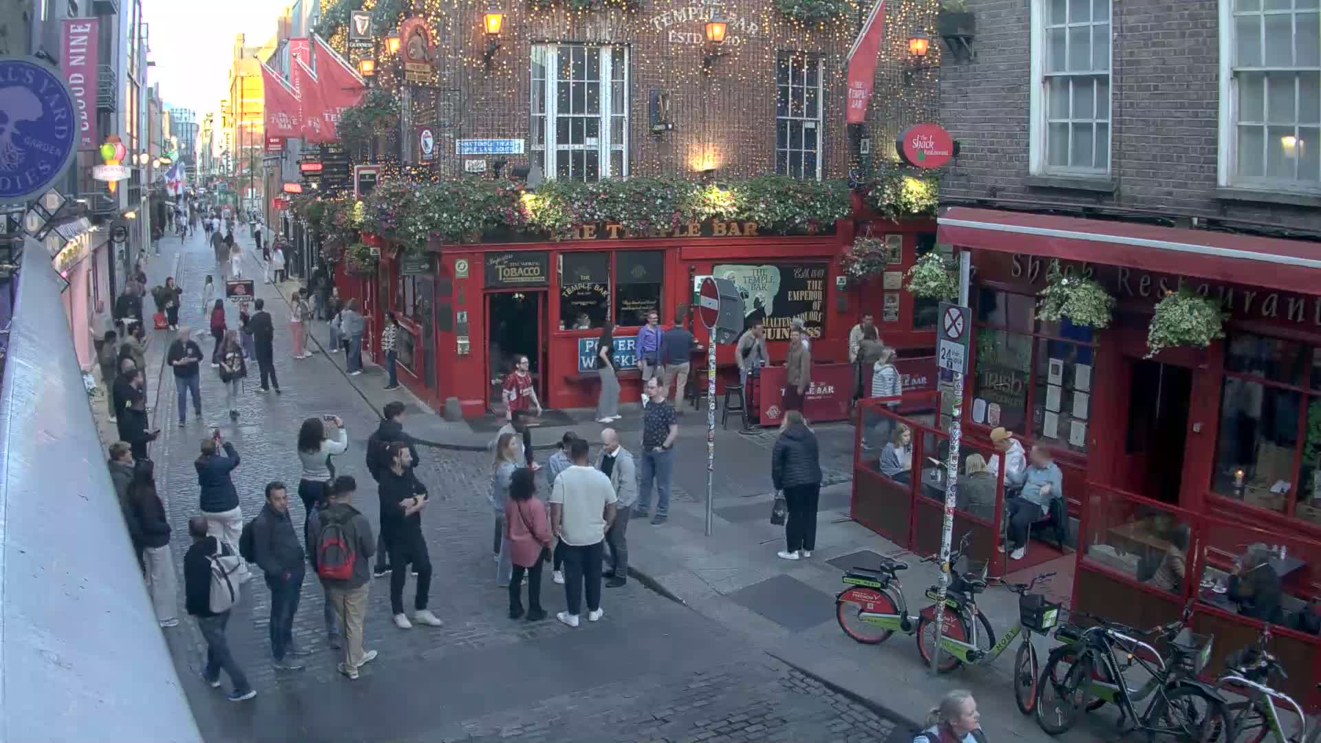 A cobblestone street, lined with red brick buildings and outdoor seating, is filled with pedestrians and several bicycles under a twilight sky.