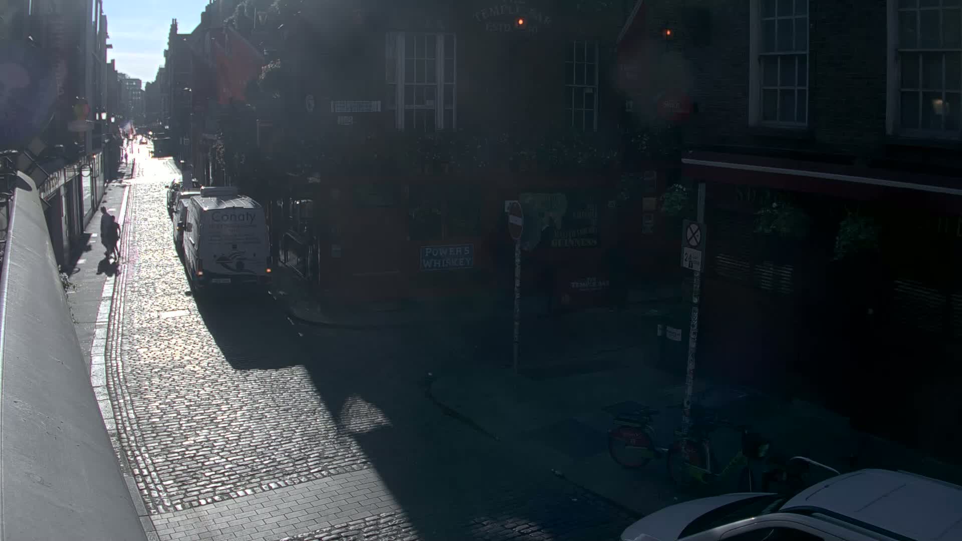 A sunny cobblestone street between buildings, with a van and pedestrians visible, and parked bicycles and cars in the lower right.