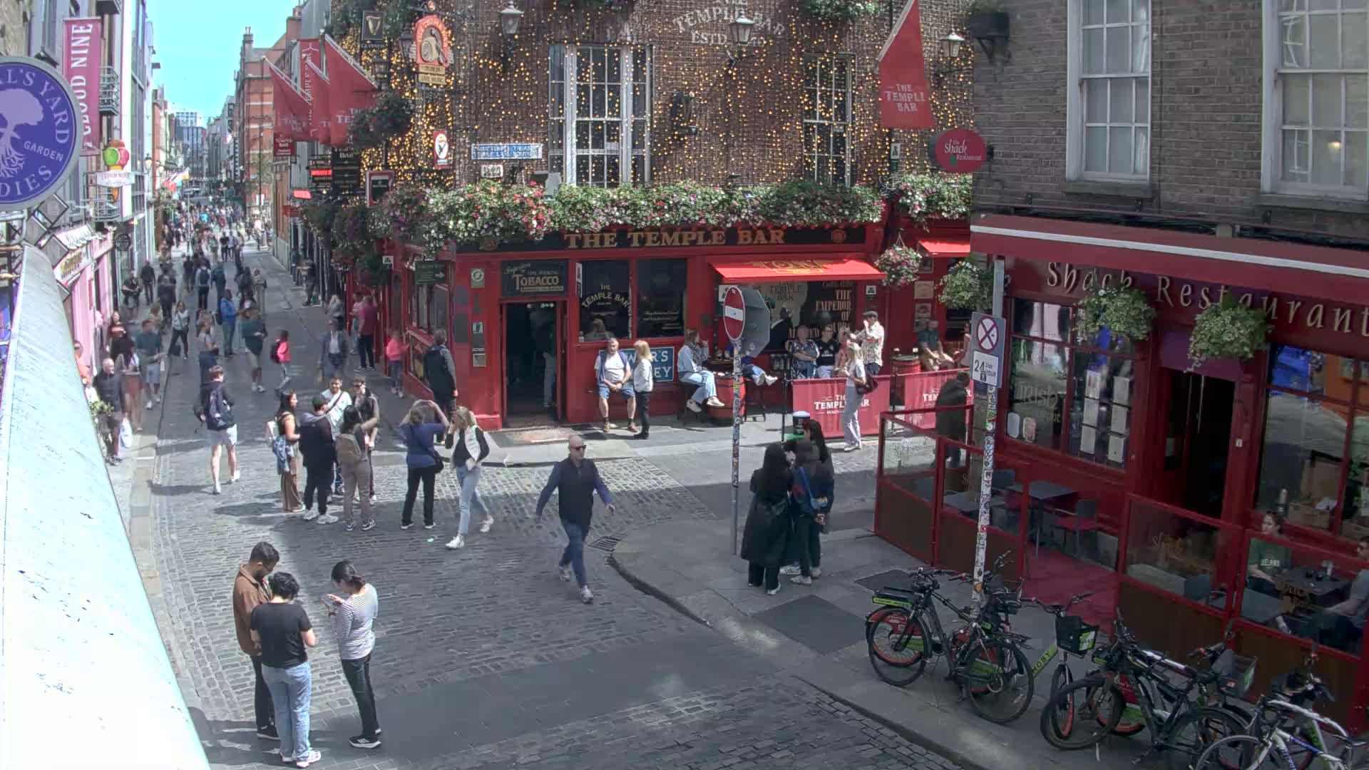A sunny day on a cobblestone street shows many people walking past red brick buildings with restaurants and pubs, including several bicycles parked along the side of the street.