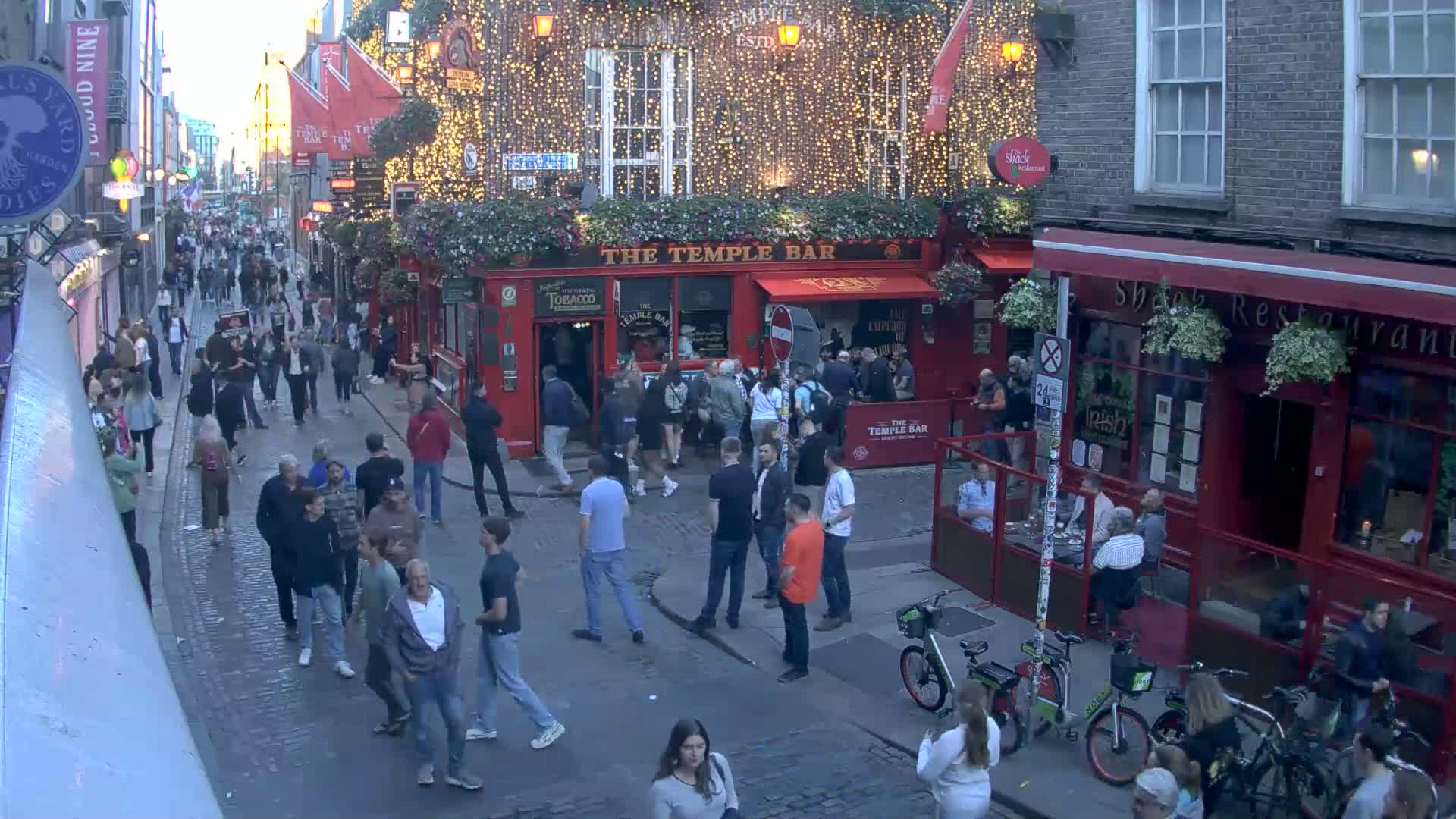 A bustling, cobblestone street at dusk is filled with pedestrians and bicycles, lined with red-brick buildings, one prominently displaying string lights and the name "The Temple Bar."