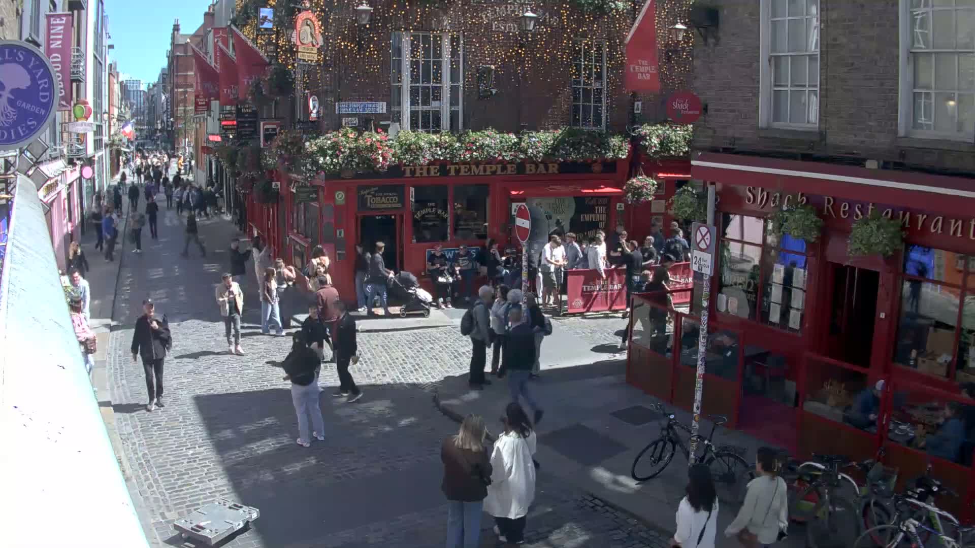 A sunny cobblestone street scene shows numerous pedestrians walking past a red-bricked pub called "The Temple Bar," and other shops and restaurants.