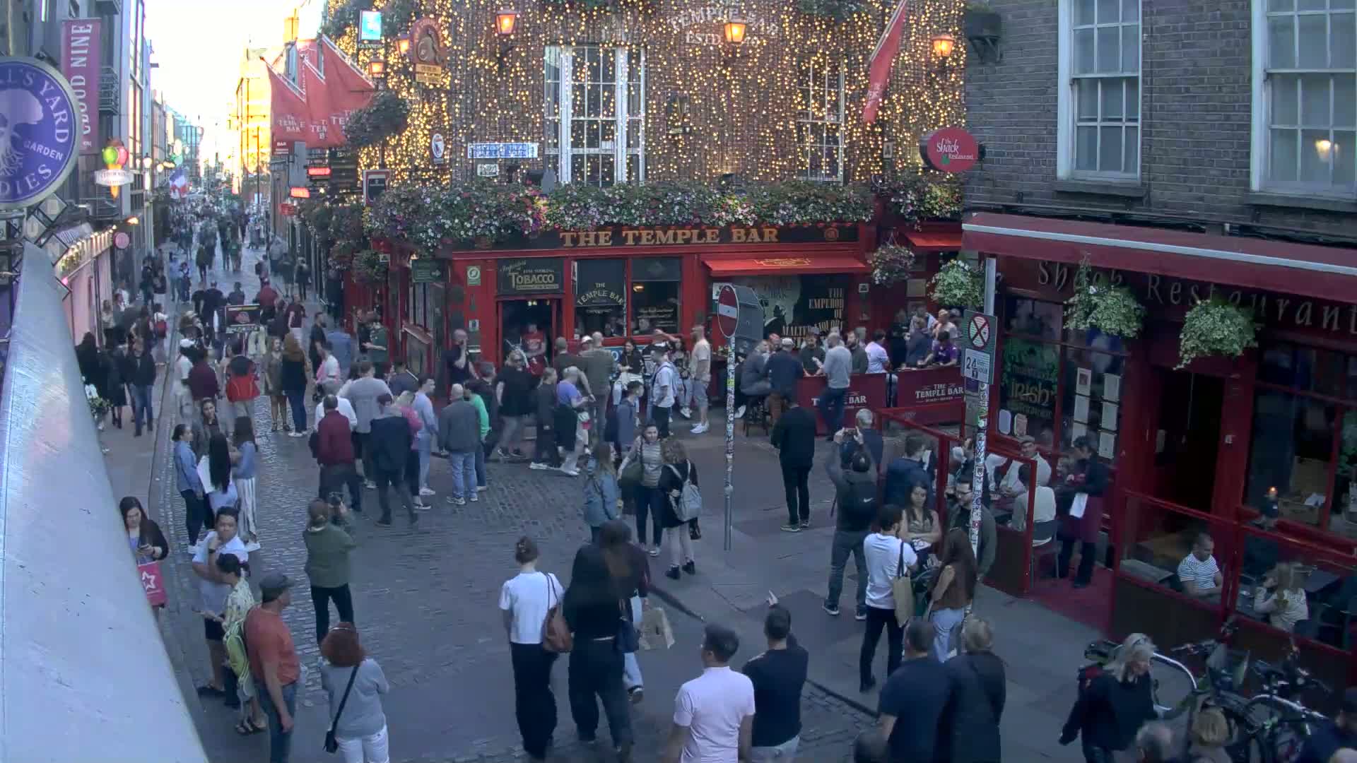 A crowded cobblestone street at dusk features a bustling pub, The Temple Bar, with patrons both inside and outside, and several nearby restaurants and shops.
