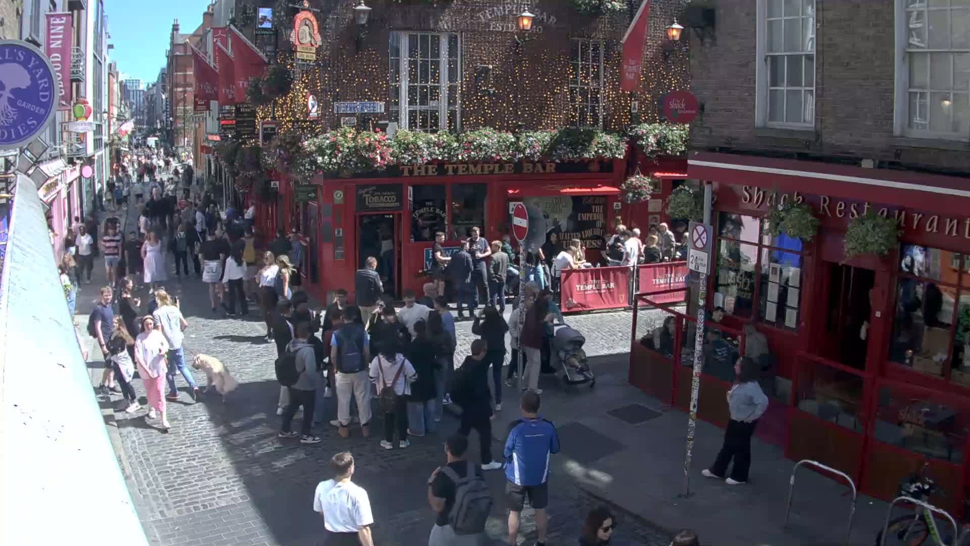 A sunny day on a cobblestone street, densely packed with pedestrians, shows a red-brick building with a pub and restaurant.