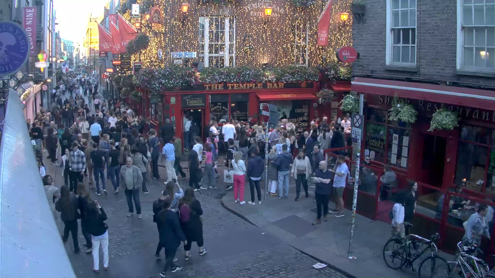A crowded cobblestone street at dusk, with numerous people gathered outside pubs and restaurants, some decorated with string lights.