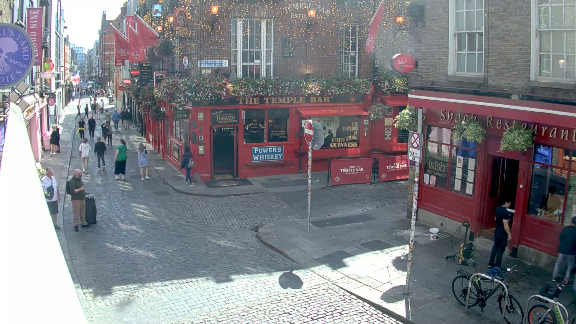 A sunny cobblestone street scene shows pedestrians walking past red brick buildings with outdoor seating and hanging plants.