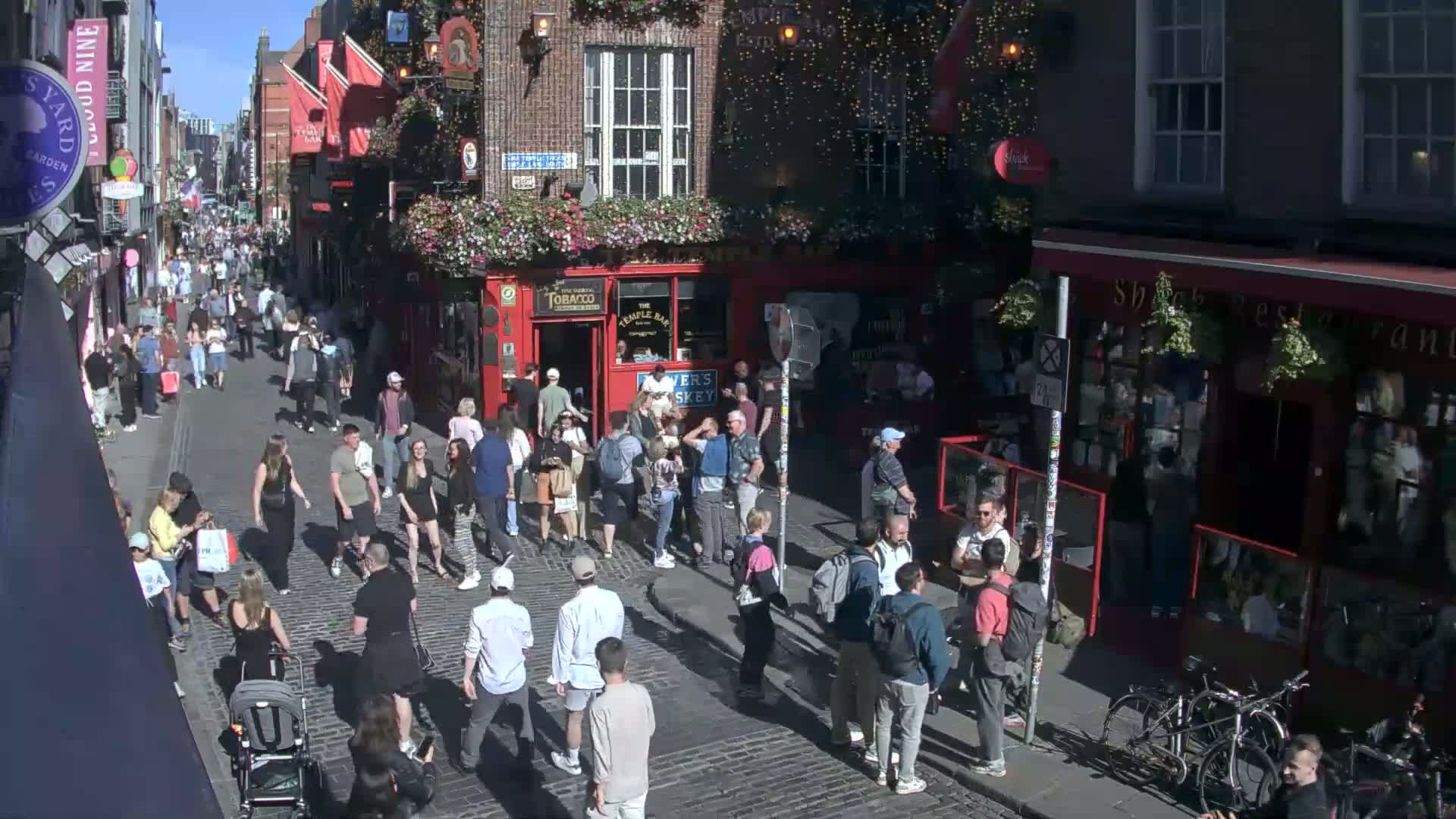 A crowded cobblestone street, under a sunny sky, features pedestrians walking past numerous shops and pubs.