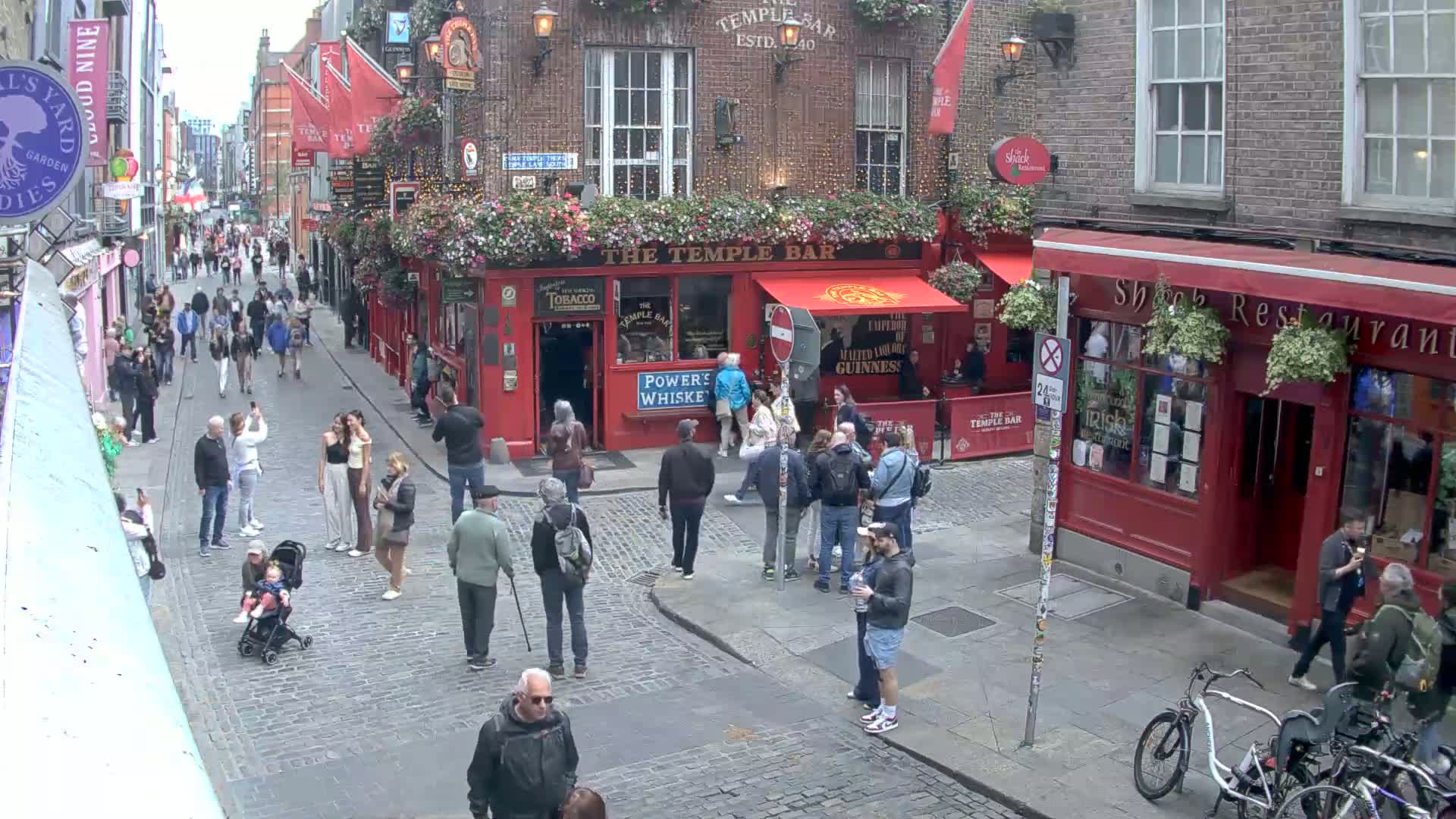 A cobblestone street is filled with pedestrians, many congregating near a red-brick pub under an overcast sky.