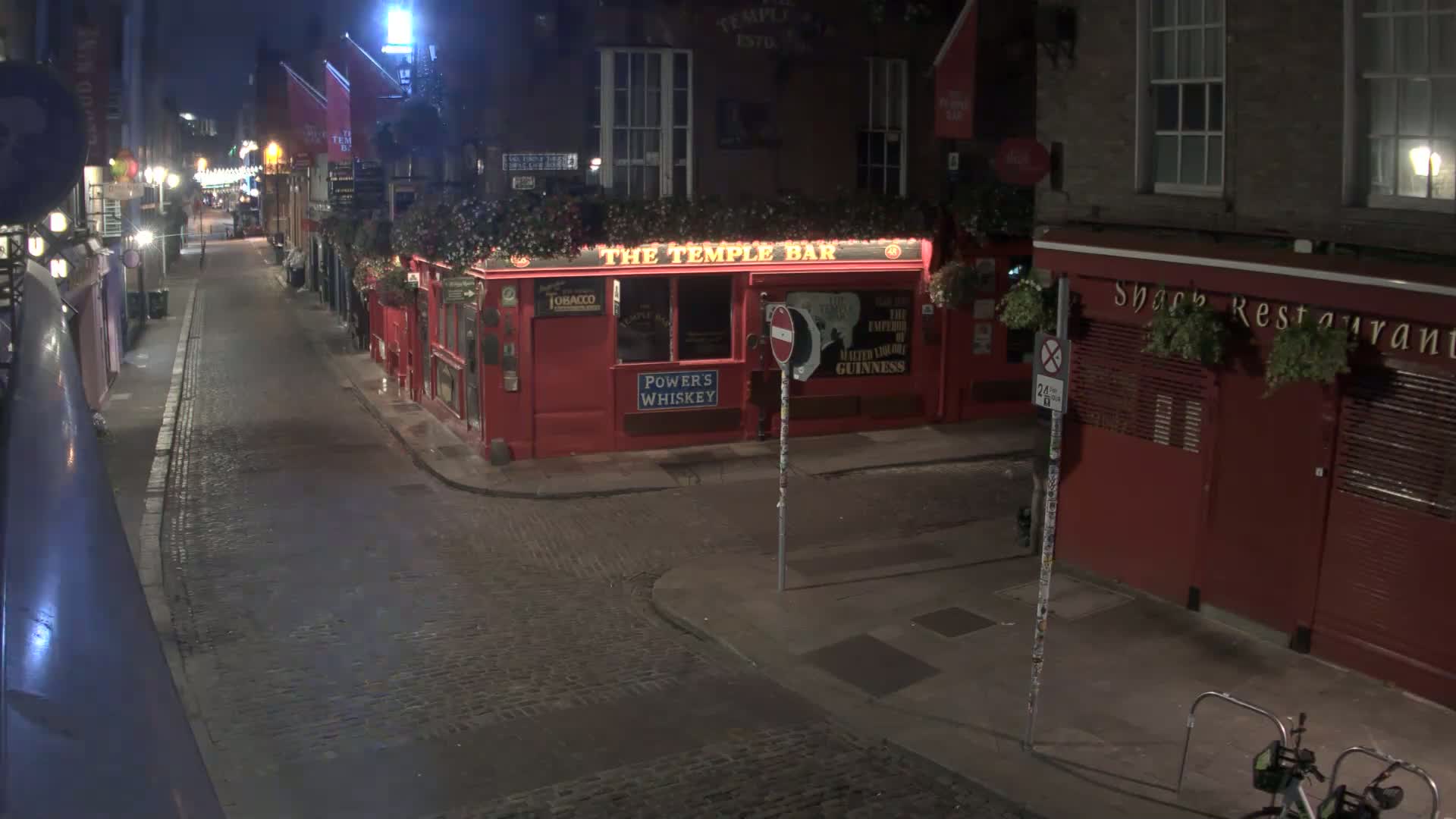 A cobblestone street at night, lined with red buildings, one of which is a brightly lit pub, is nearly deserted under dark skies.