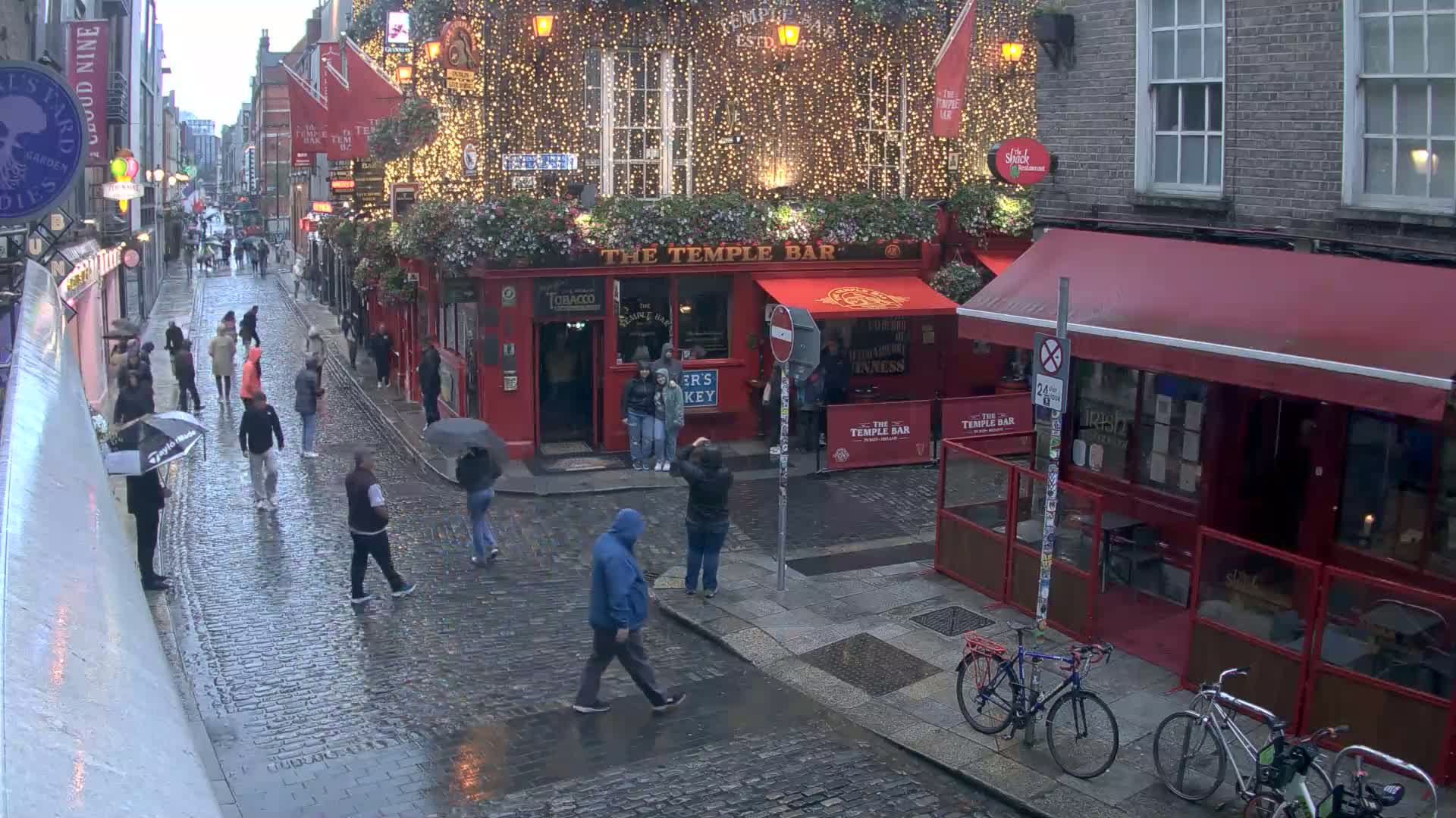 A rain-slicked cobblestone street, lined with buildings and pedestrians, features a brightly lit pub decorated with string lights.