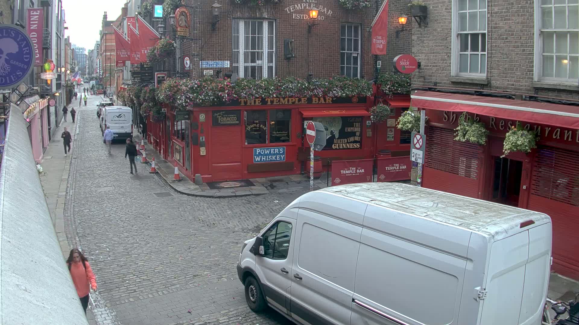 A cobblestone street scene on an overcast day shows a red-brick building with a pub, people walking, and a white van parked.
