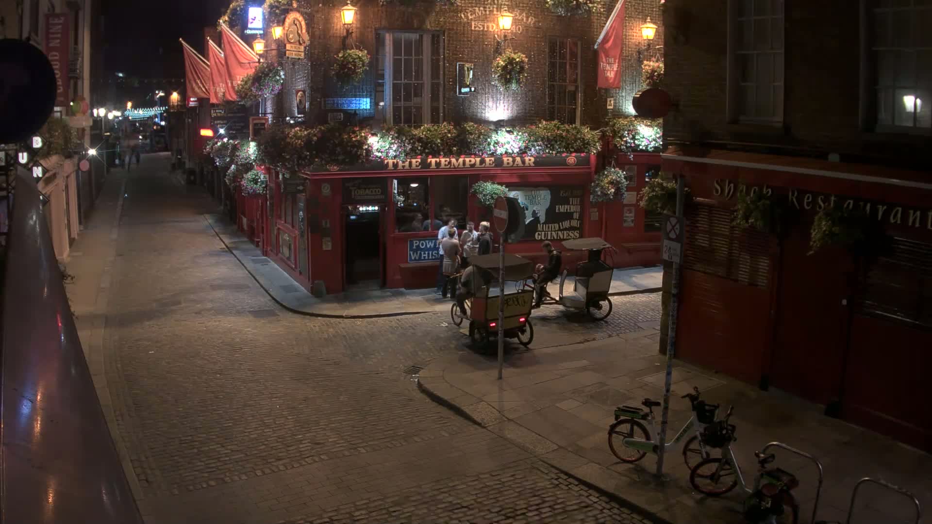 A cobblestone street at night is illuminated, showing a pub with people outside and rickshaws parked nearby, along with parked bicycles.