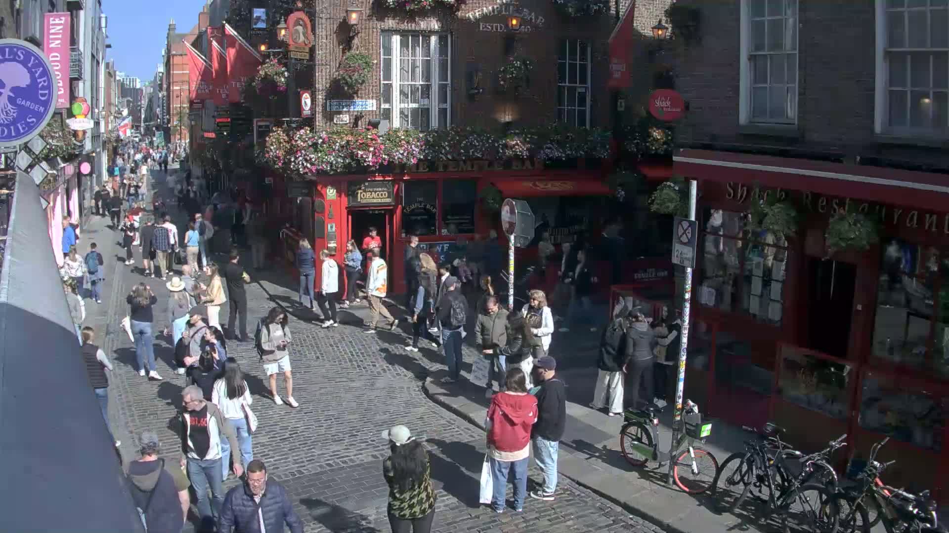 A sunny cobblestone street is crowded with pedestrians, some walking and others standing near shops and a pub.