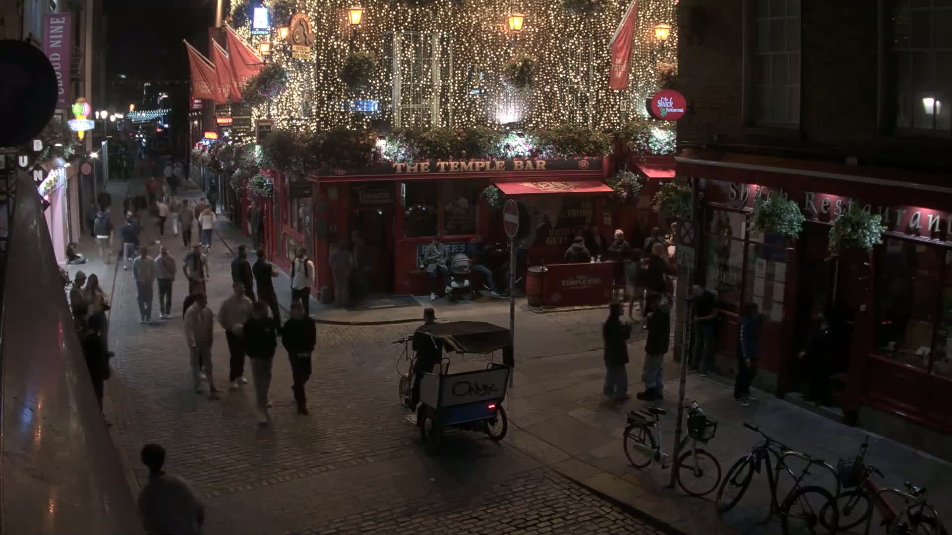 A nighttime street scene shows a cobblestone street with pedestrians, a rickshaw, bicycles, and several illuminated buildings, including a pub decorated with strings of lights.