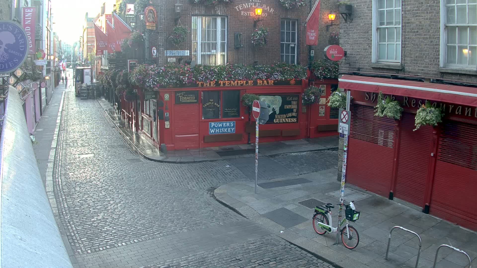 A cobblestone street, wet from recent rain, shows red brick buildings with hanging flower baskets and a parked bicycle in front of a pub.