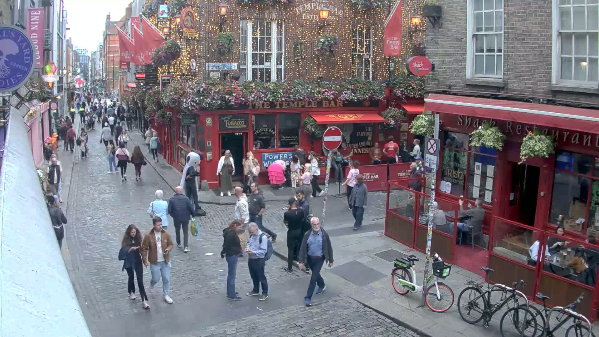 A bustling cobblestone street scene on an overcast day shows many pedestrians walking past numerous pubs and restaurants, some with outdoor seating and string lights.