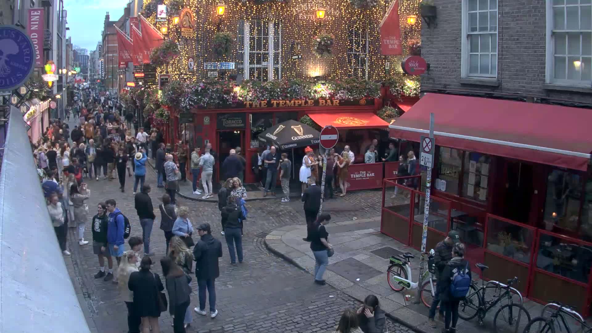 A crowded cobblestone street at dusk features a bustling pub decorated with string lights, and several people are walking around, with bicycles parked along the side.