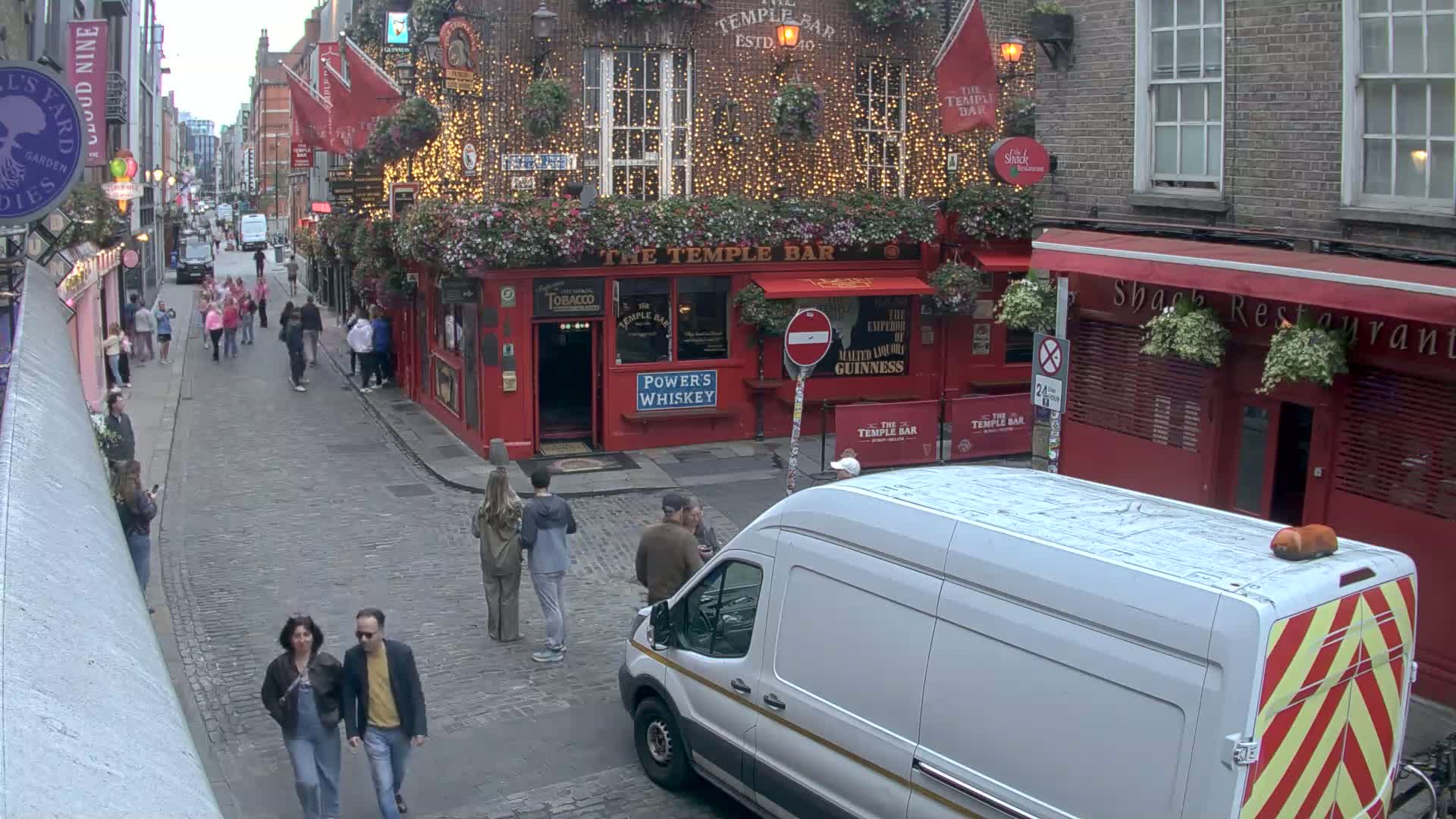 A cobblestone street scene shows pedestrians walking past a red brick building with a pub called "The Temple Bar" decorated with string lights and hanging flower baskets, under overcast skies.