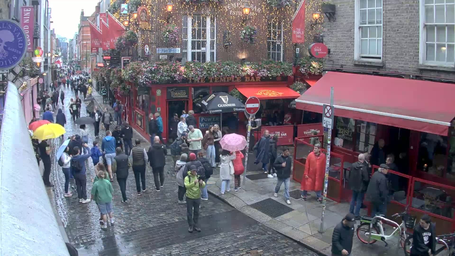A rain-soaked cobblestone street is crowded with pedestrians carrying umbrellas, passing by red-awninged pubs and shops.