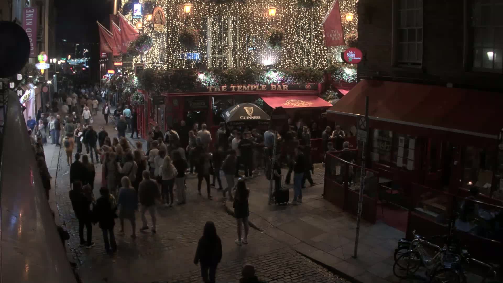 A nighttime street scene shows a crowded area outside a brightly lit pub decorated with string lights, with people milling about on a cobblestone street.