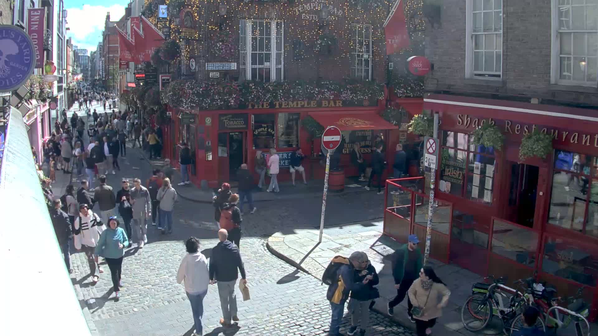 A sunny day on a cobblestone street in a city, crowded with pedestrians, features red brick buildings with pubs and restaurants.