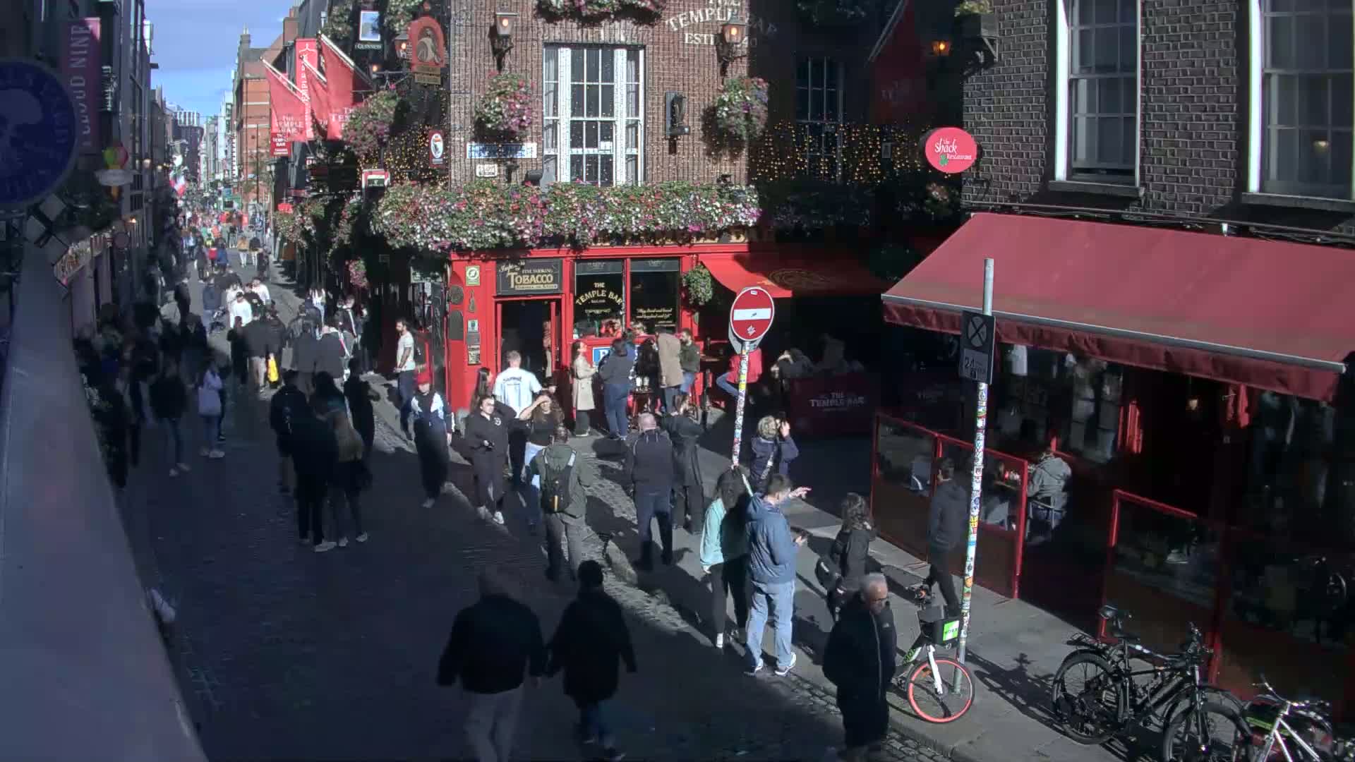 A sunny day on a cobblestone street with many people walking past shops and pubs, some of which have red awnings.