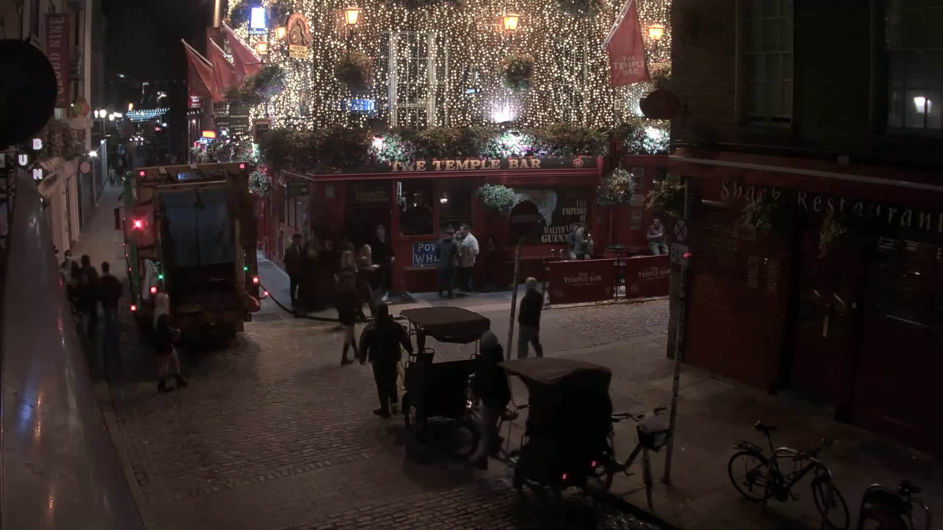 At night, a street scene shows a garbage truck and pedestrians near a brightly lit pub decorated with string lights, and a few rickshaws and bicycles are parked on the cobblestone street.
